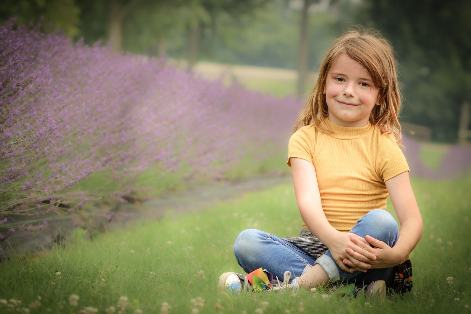 Child in yellow shirt sits cross-legged on grass near blooming lavender flowers.