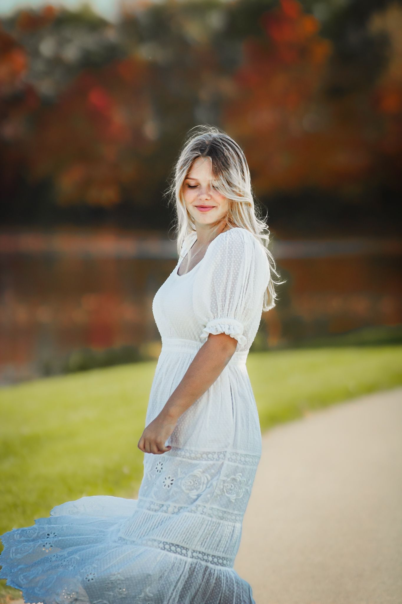 Person in flowing white dress poses on path with autumn colors in background.