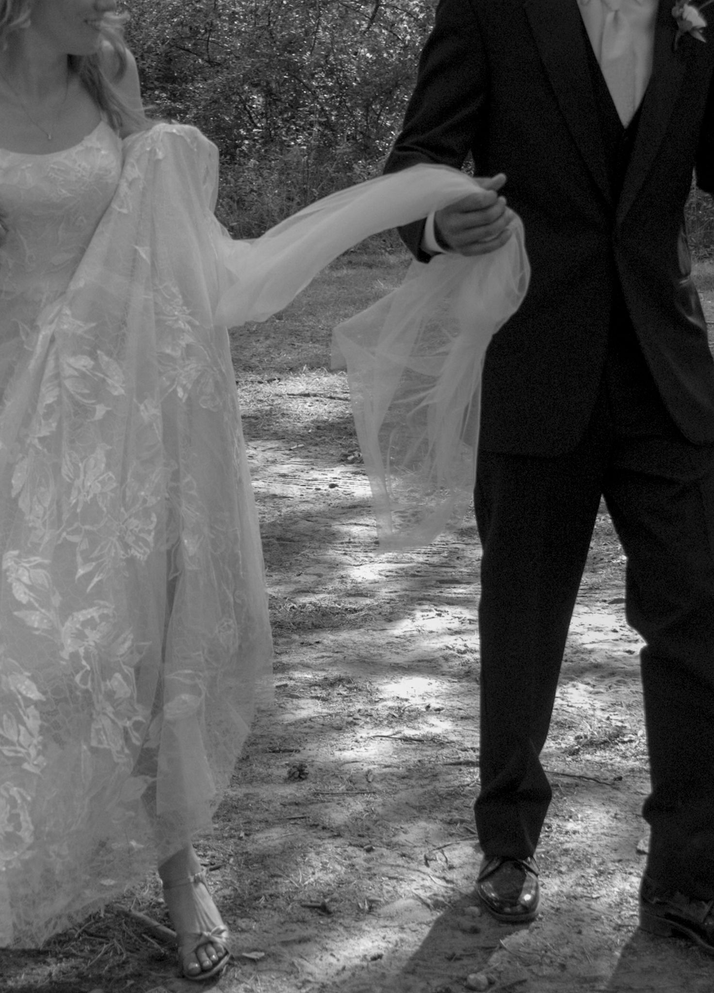 A couple in wedding attire walking on a sunlit forest path captured in black and white.