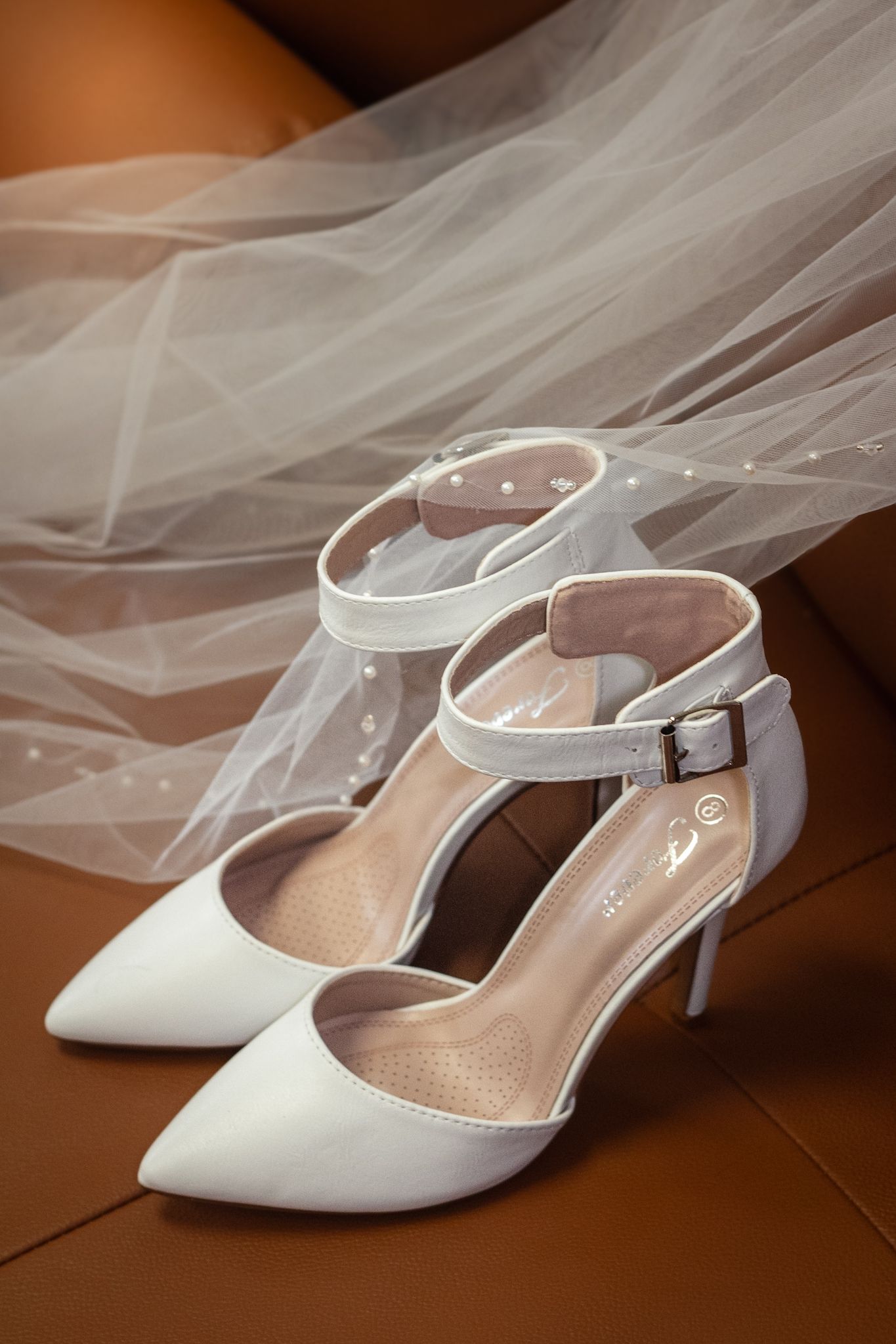 Elegant white bridal shoes with ankle straps displayed with flowing veil on brown leather surface.