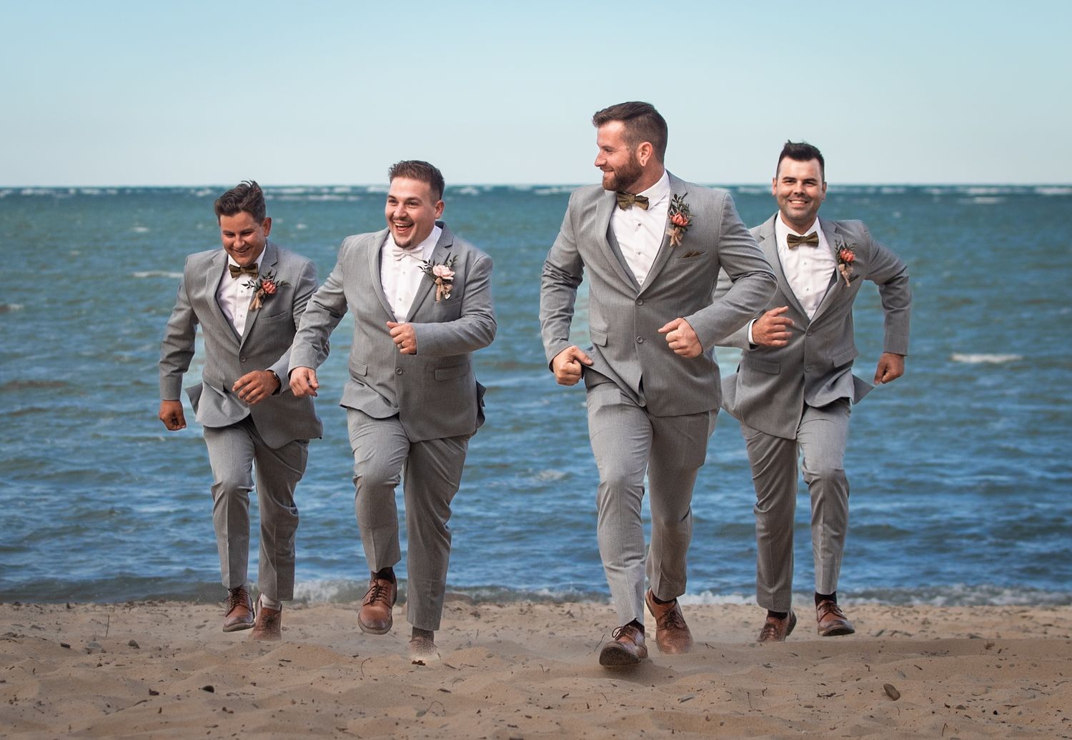 Groomsmen in matching gray suits run playfully along a beach with ocean waves in the background.