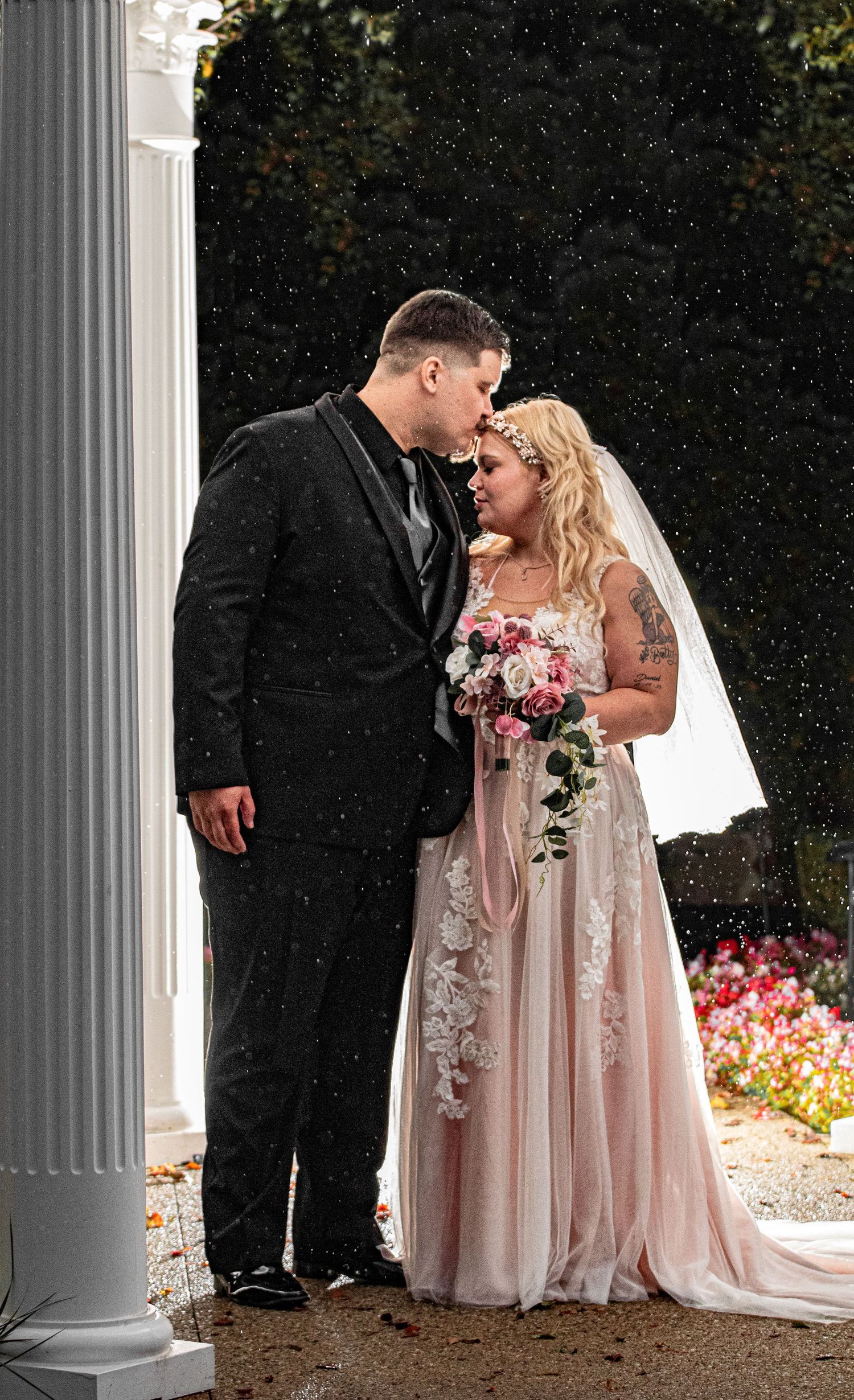 Wedding couple shares intimate moment between classic white columns with pink floral arrangements.
