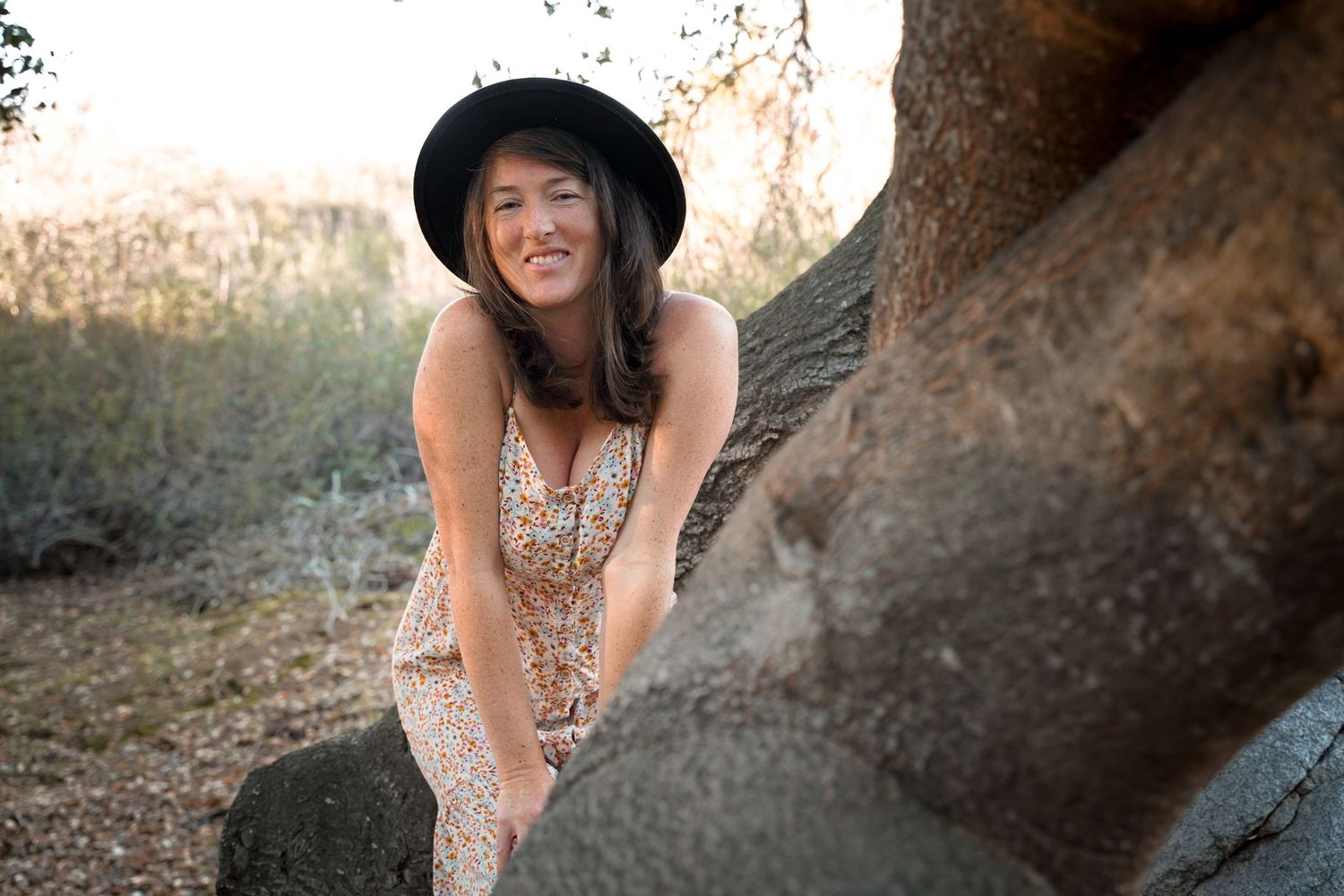 Person in hat and dress smiling while leaning against large tree trunk at sunset.
