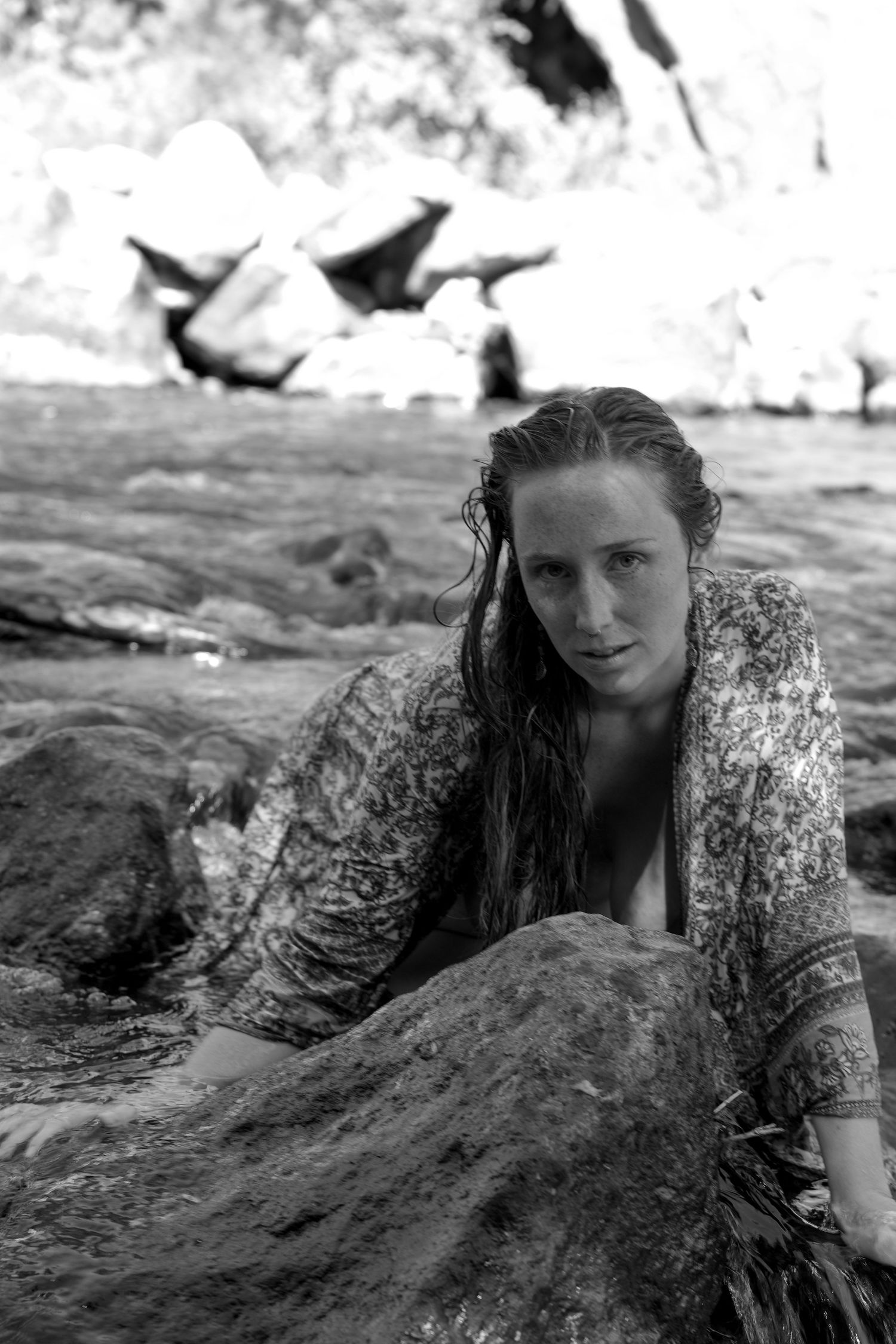 A black and white portrait of someone resting on coastal rocks while wearing a patterned top.