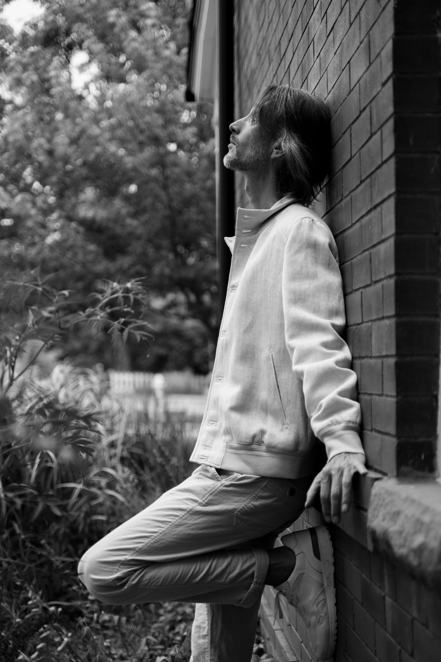A person in a white jacket leans against a brick wall outdoors in a contemplative black and white photograph.