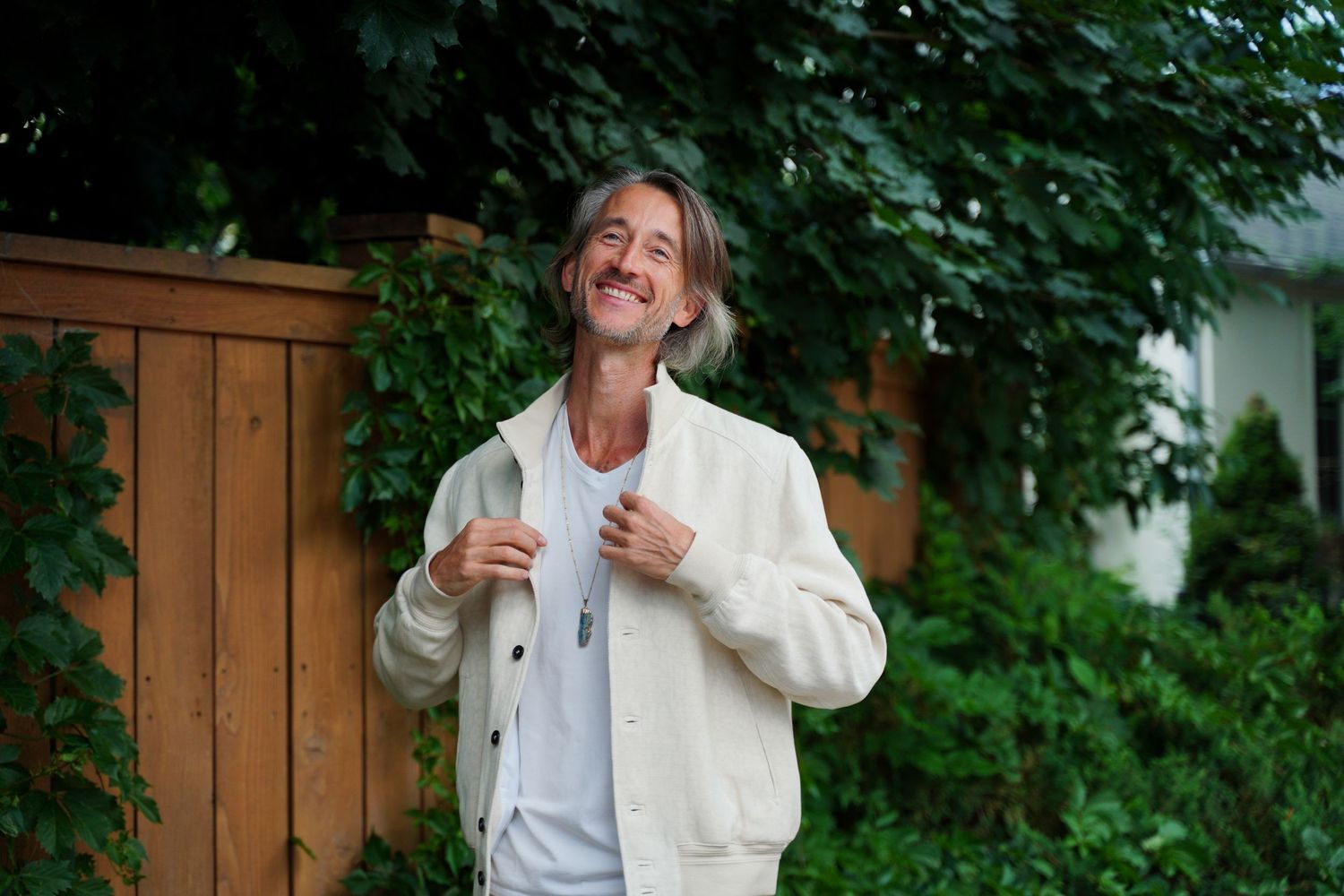 Smiling person wearing a white jacket stands outdoors in front of wooden fence and green foliage.