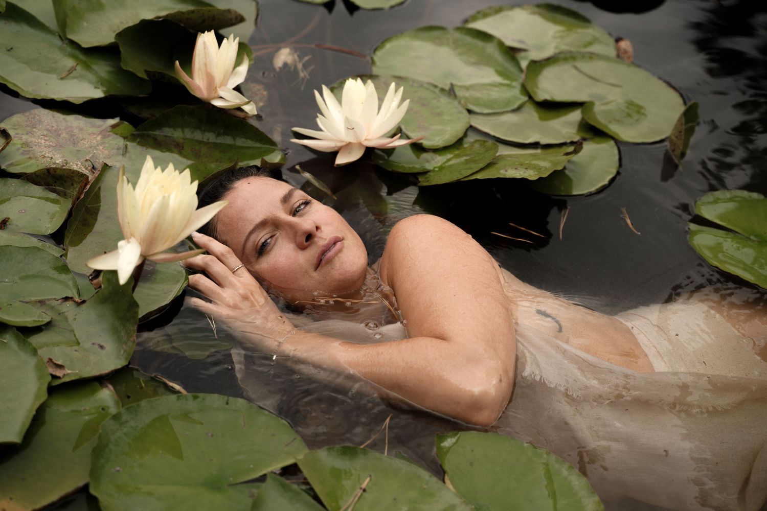 A person poses artistically in dark water among blooming white water lilies and lily pads in a moody nature scene.