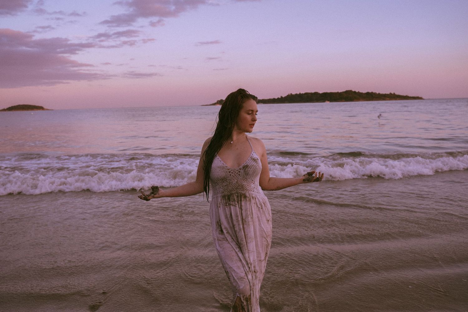 Figure in flowing dress stands in ocean waves during purple sunset.