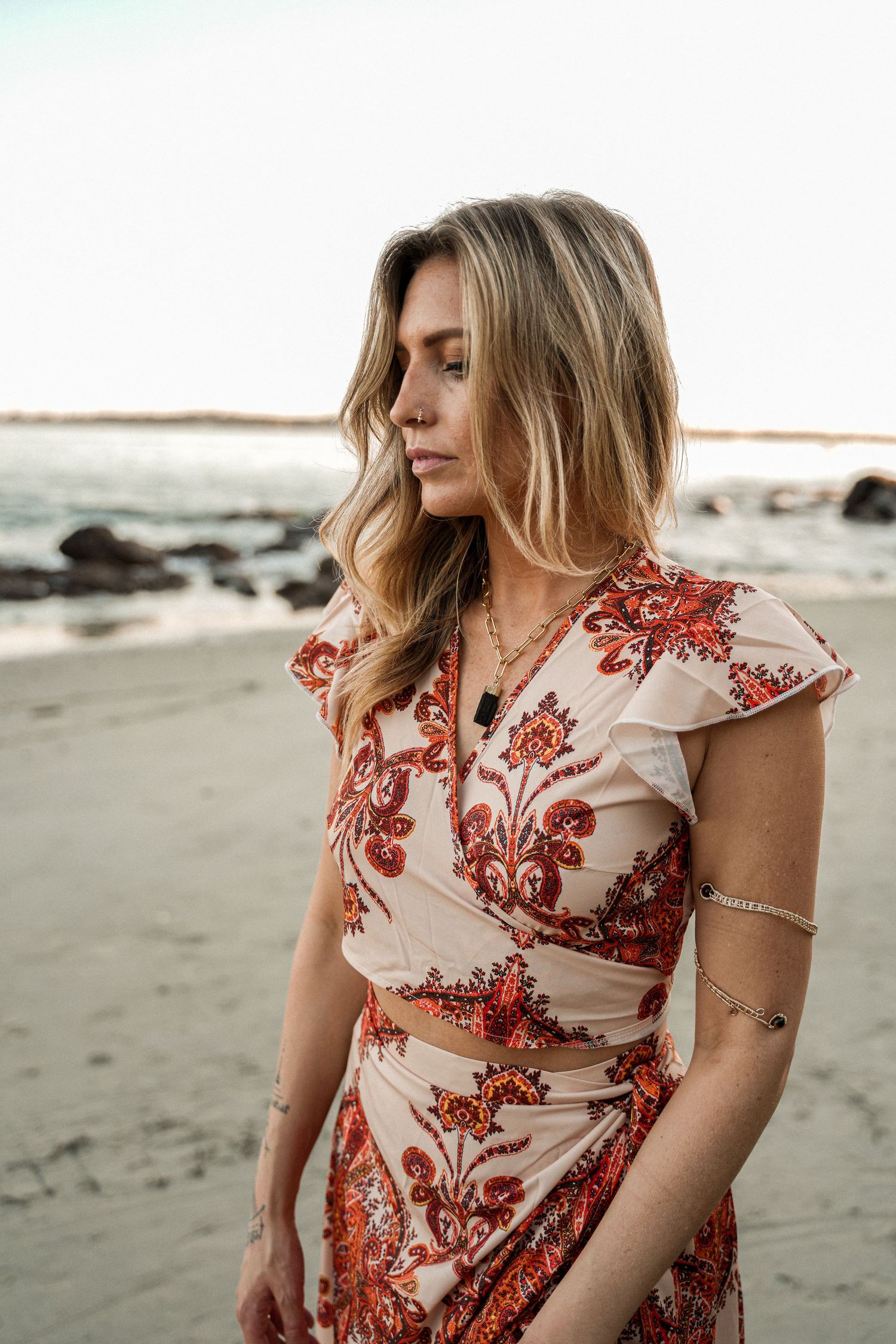 A floral print two-piece outfit photographed on a beach at sunset with rocks in the background.