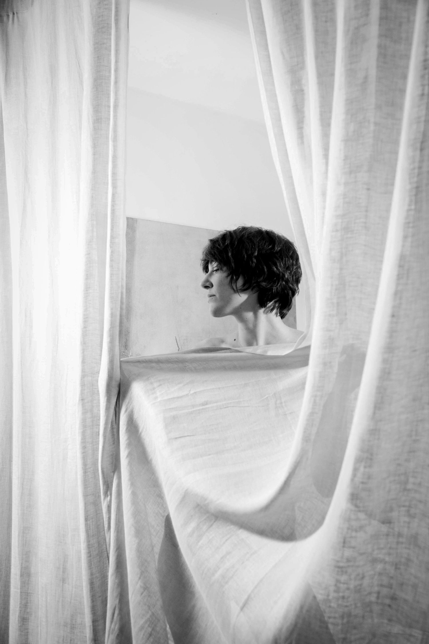 Black and white photo of figure partially concealed behind flowing white curtains.