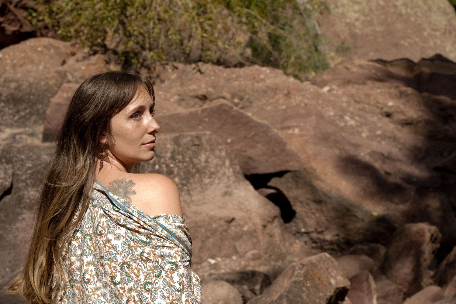 A person in a floral off-shoulder top poses against rocky red desert formations on a sunny day.