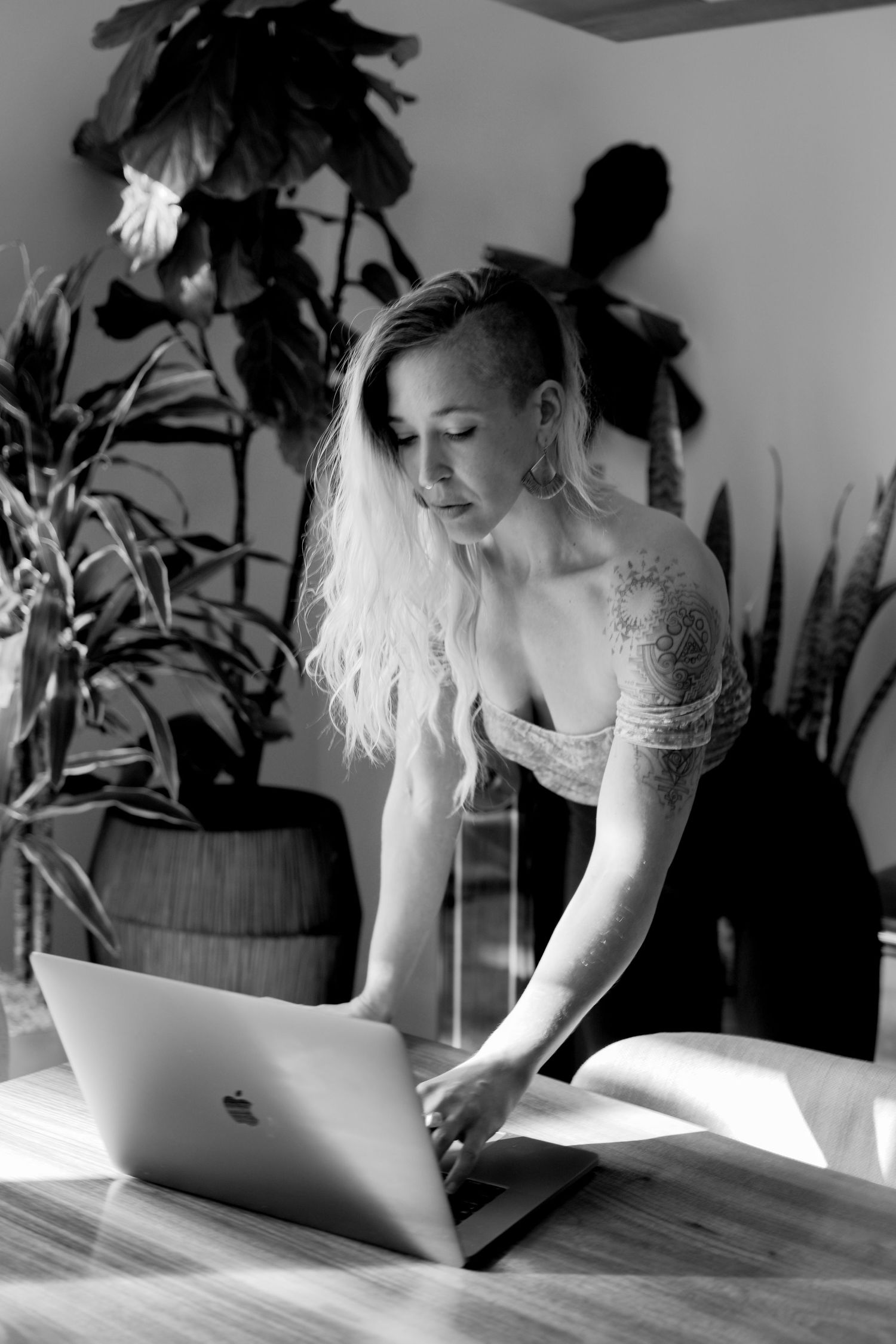 A black and white photo of someone working on a MacBook laptop in a plant-filled room.