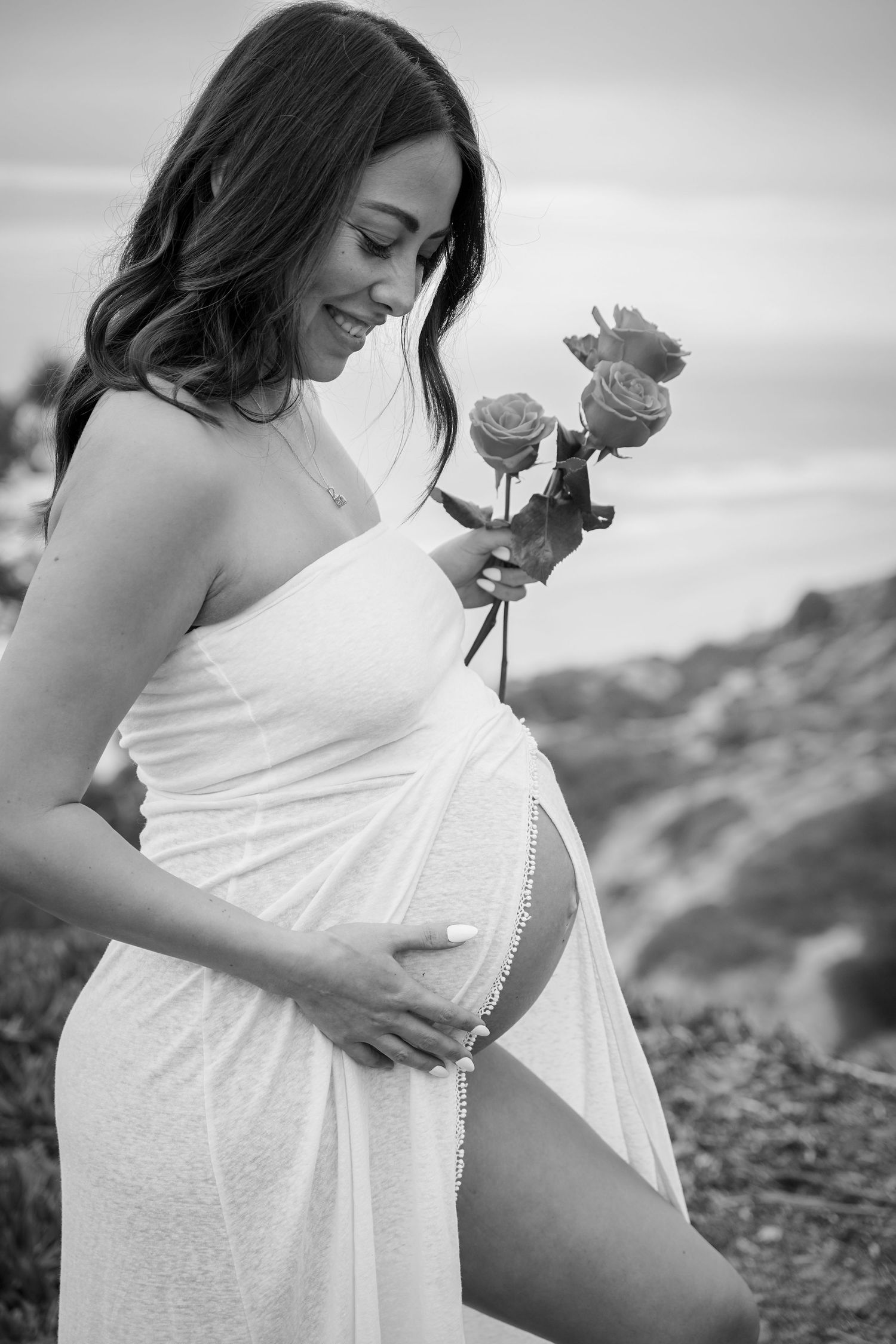 Expectant mother in white dress smiles while holding roses against coastal backdrop.