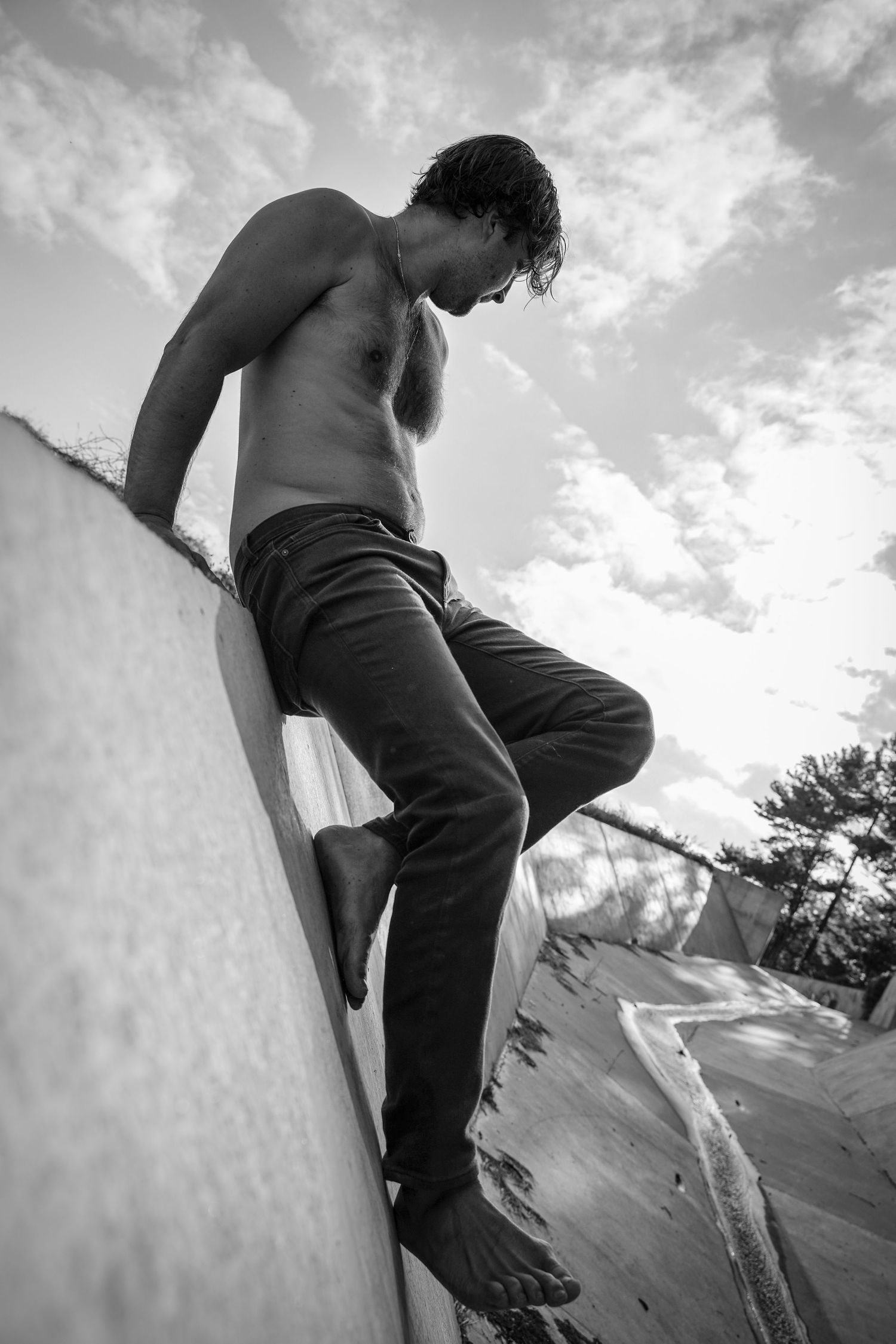 Black and white low angle photo of shirtless figure climbing concrete wall against cloudy sky.