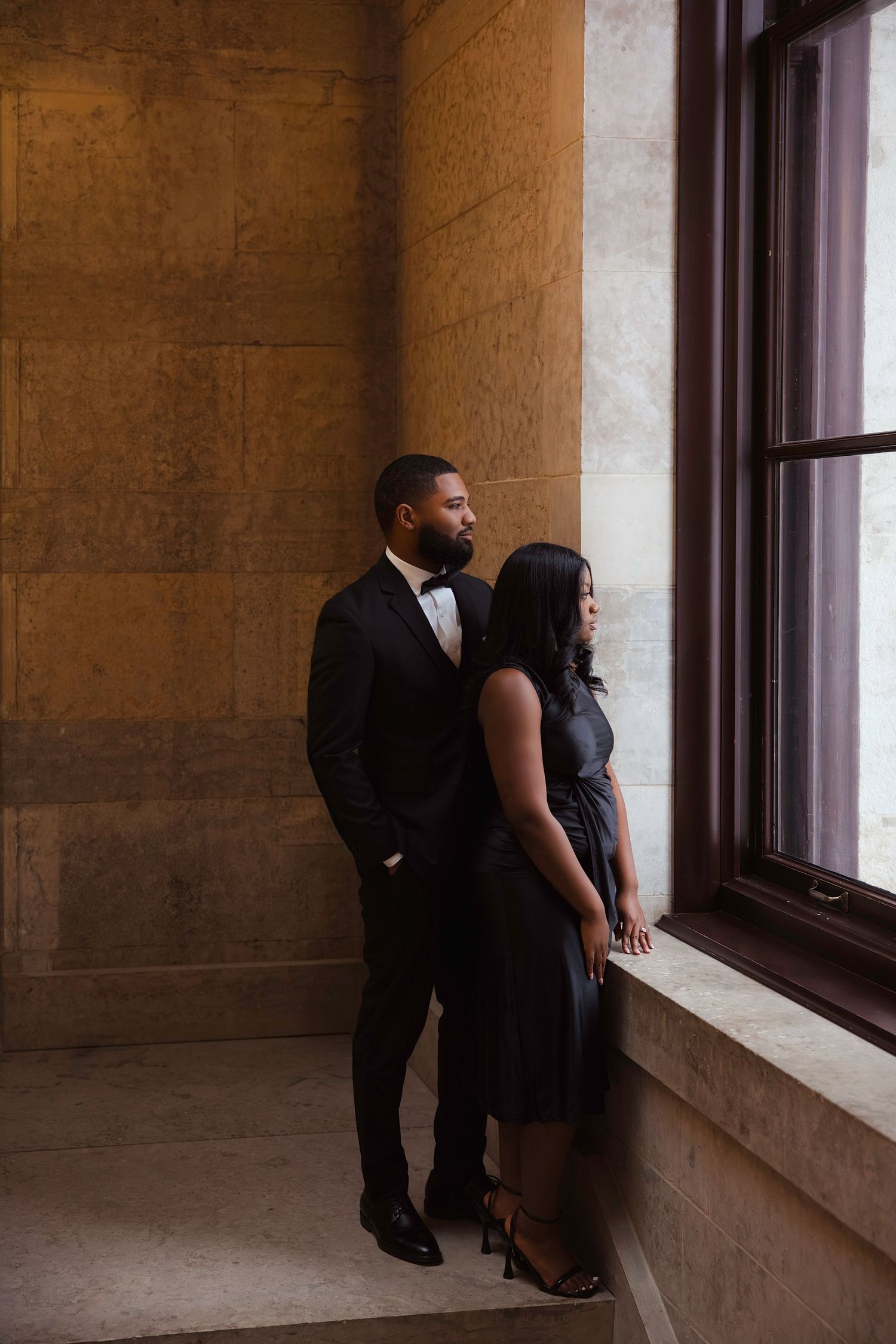 A couple in formal black attire stands near a window in an elegant stone building interior.