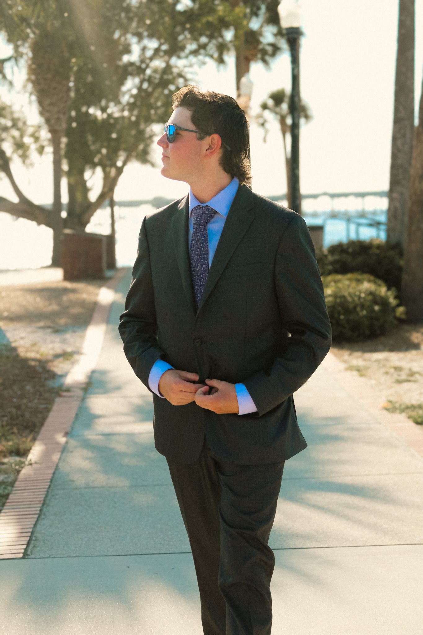 Portrait of a young man during his Prom walking along a sunlit path near the water. He wears a tailored dark suit with a blue dress shirt.