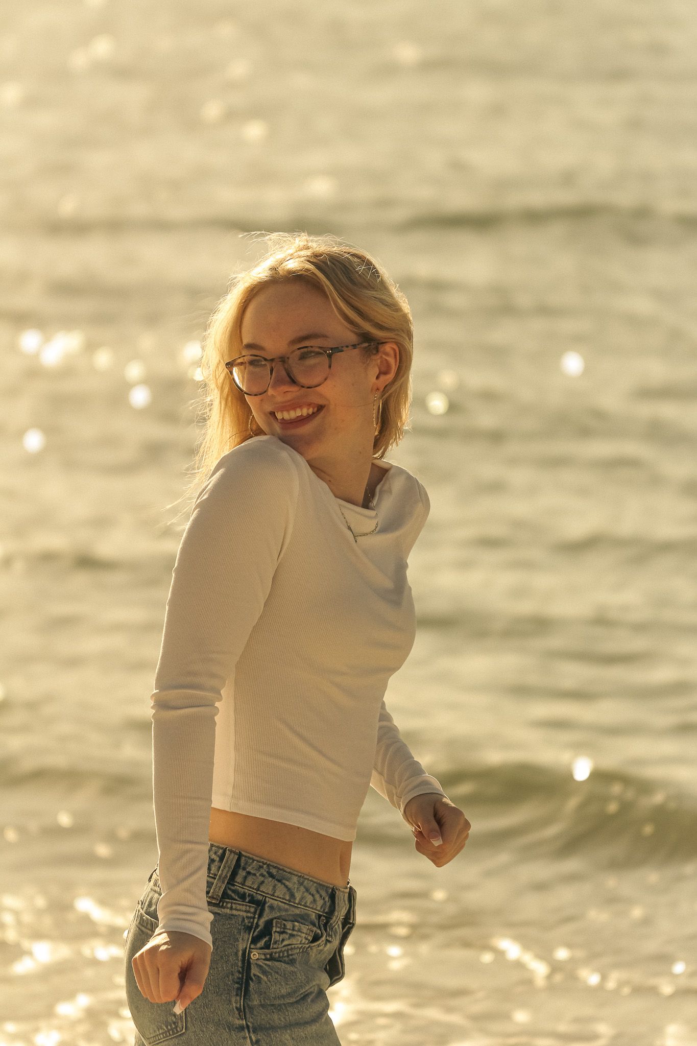 A candid, sunlit beach portrait of a young woman standing by the shoreline. She’s turned slightly to the side, smiling warmly.