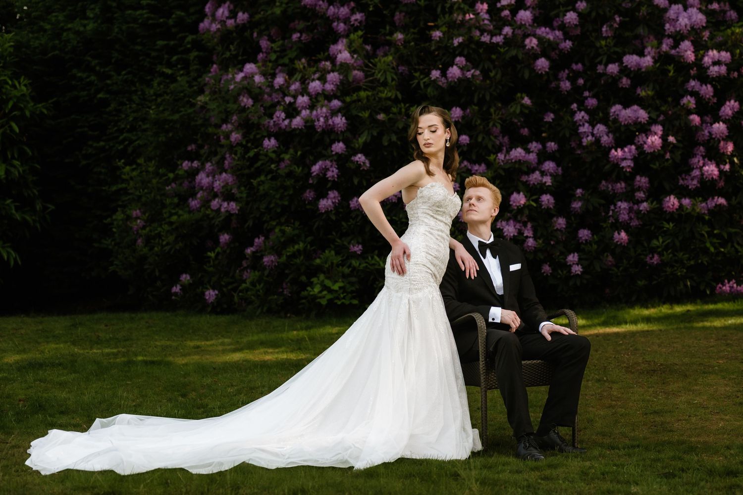 Elegant outdoor wedding portrait of a bride standing beside her seated groom in a garden with purple flowers.
