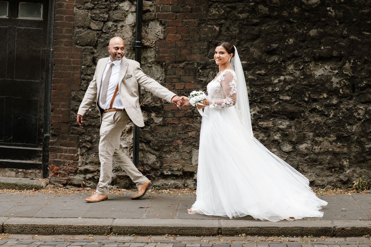 Bride and groom holding hands while walking beside a rustic stone wall on their wedding day.