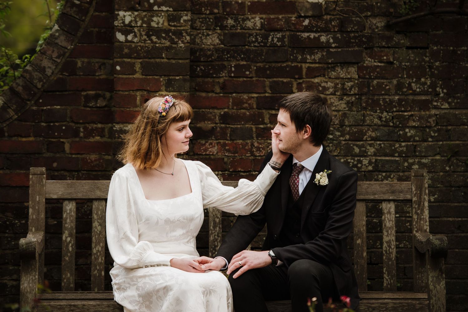 Bride and groom sitting together on a wooden bench, sharing a tender moment against a rustic brick wall.