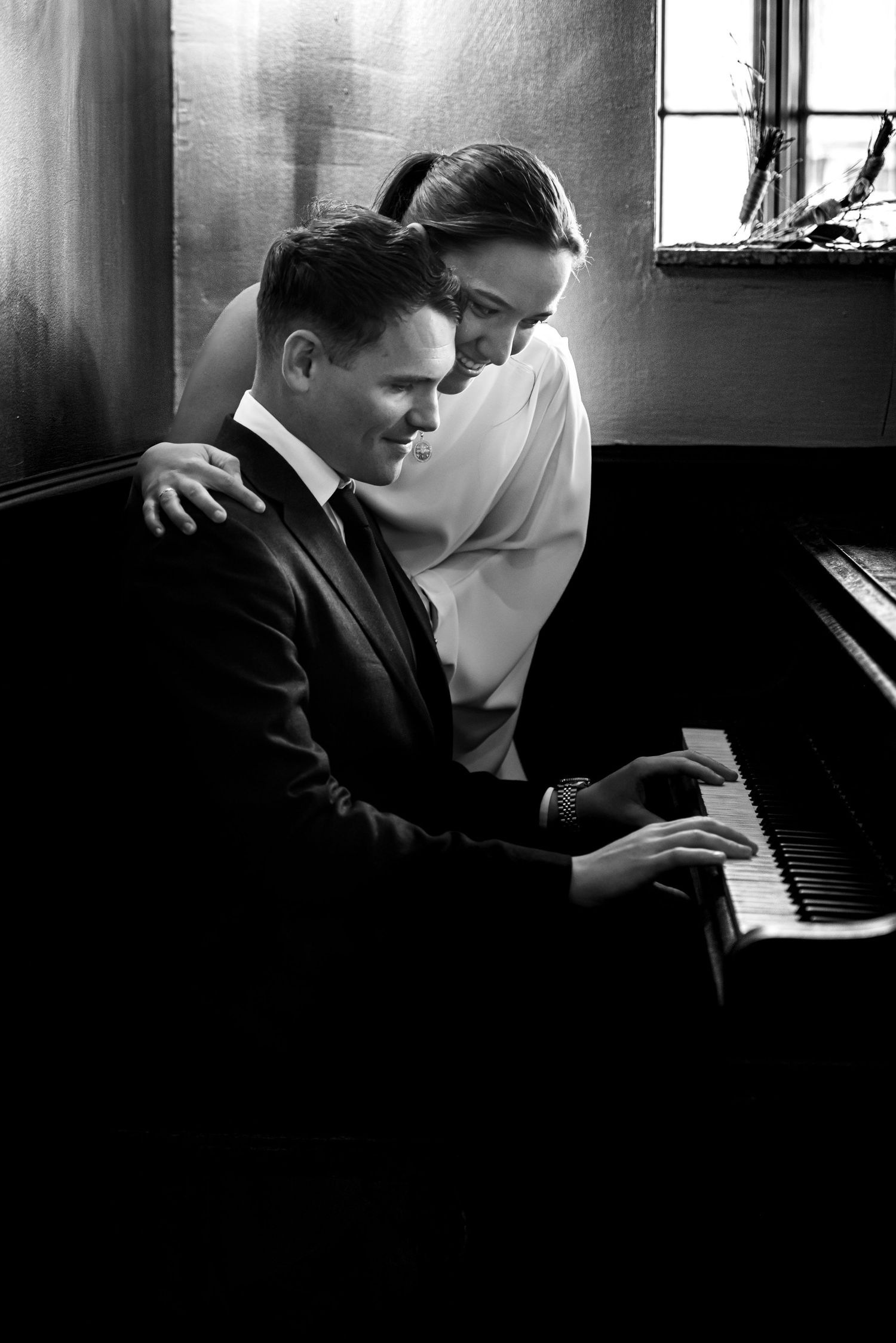 Black and white photograph of a bride embracing the groom as he plays the piano.