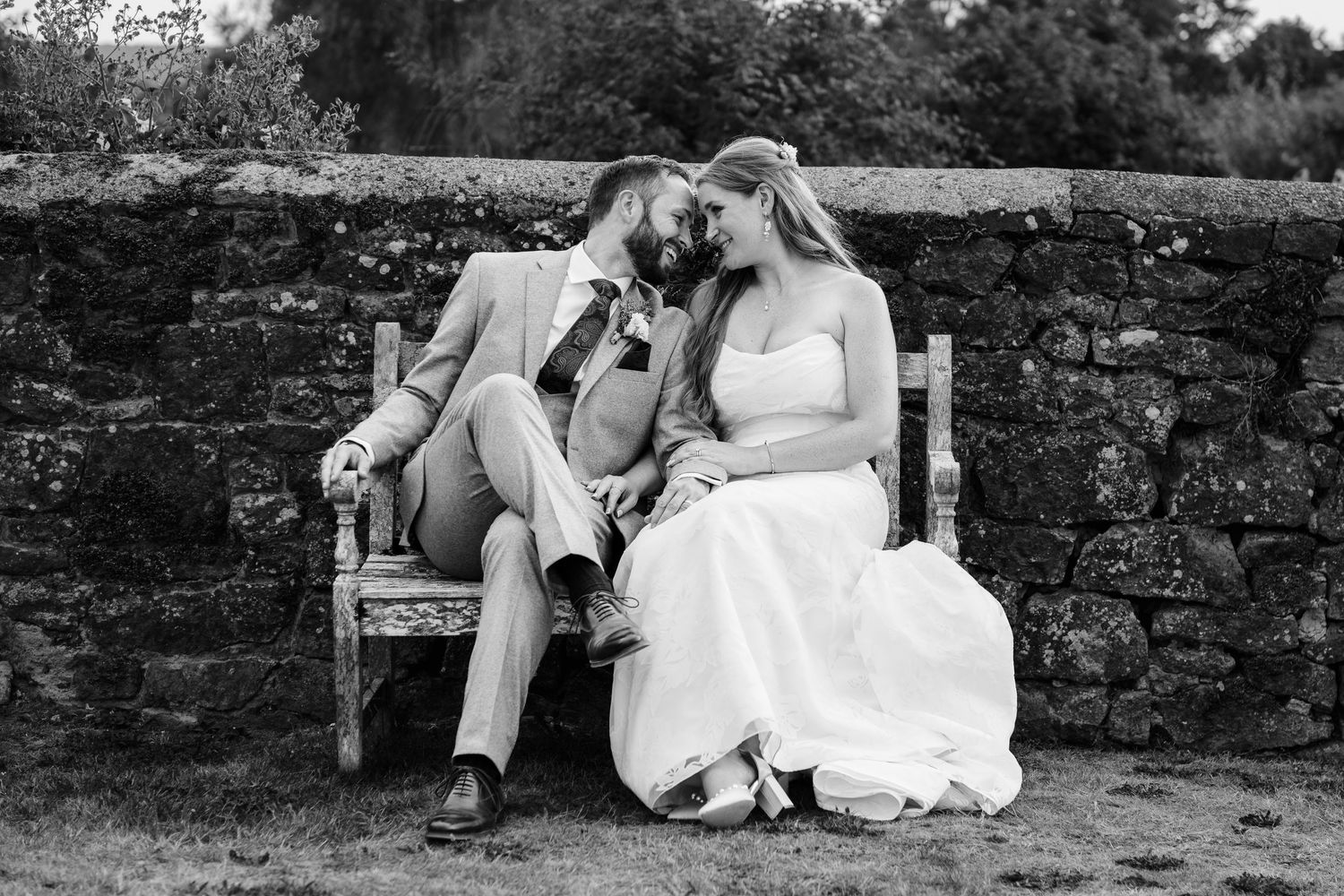 Black and white wedding portrait of a bride and groom sitting closely together on a bench beside a stone wall.