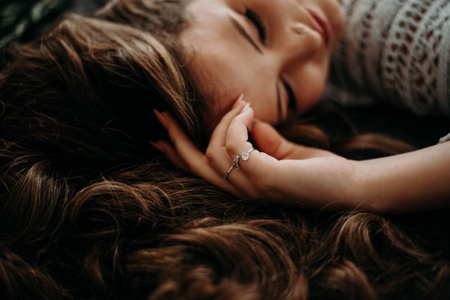 A smiling woman with long brown hair lies down, wearing a diamond ring, eyes closed in joy.