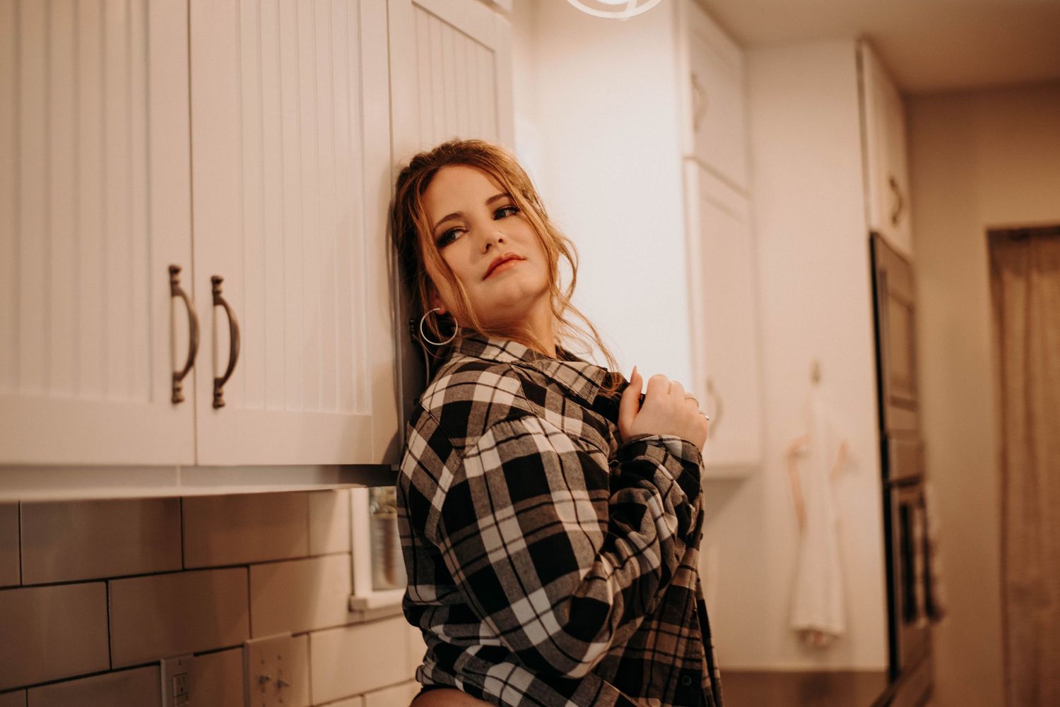Young woman in plaid shirt leaning against white kitchen cabinets with a confident pose.