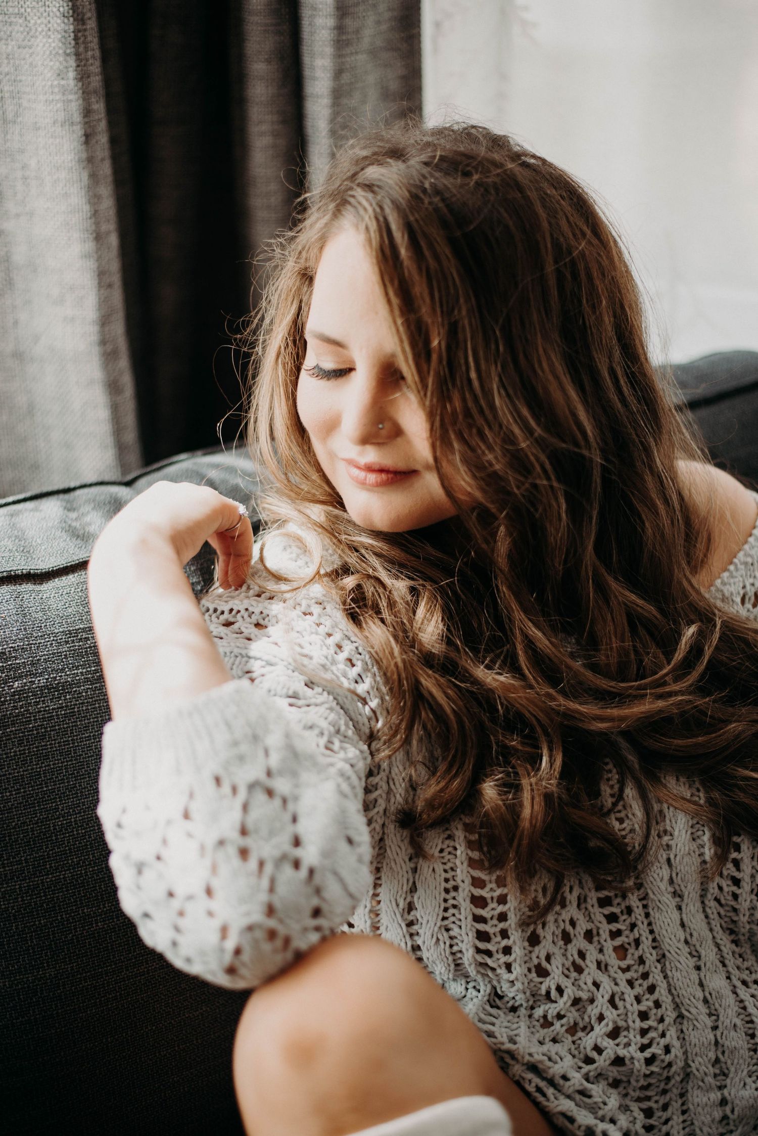 Young woman with long wavy brown hair looking at a watch while sitting on a gray sofa.