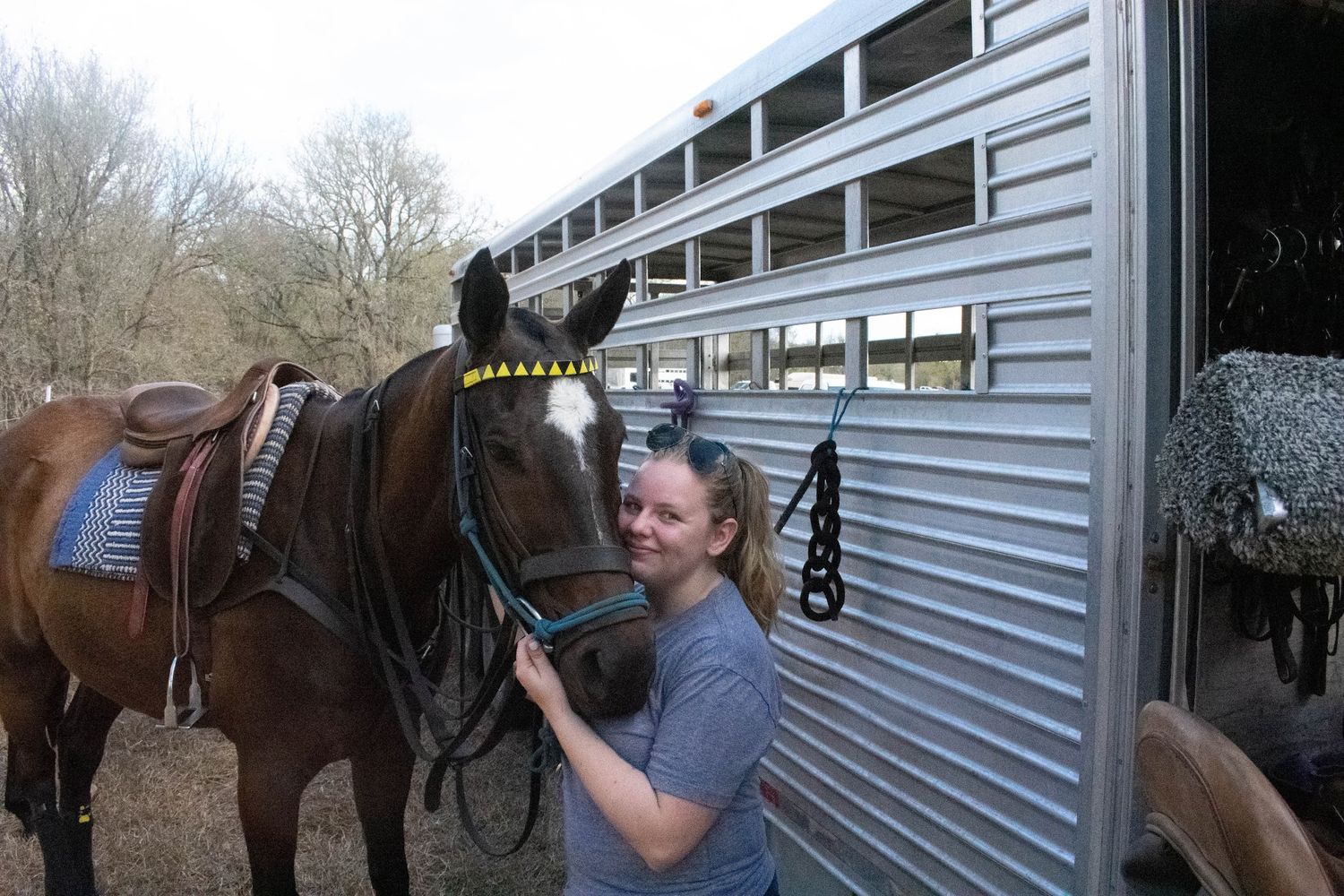 Smiling woman standing beside a saddled brown horse near a silver horse trailer outdoors.