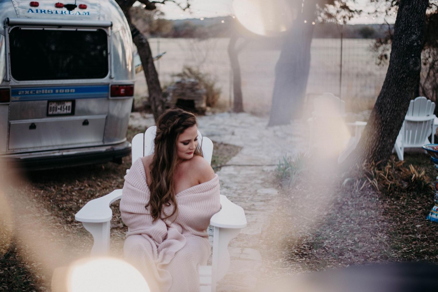 Woman in pink off-shoulder dress sits in white chair near vintage Airstream trailer outdoors in soft golden light.