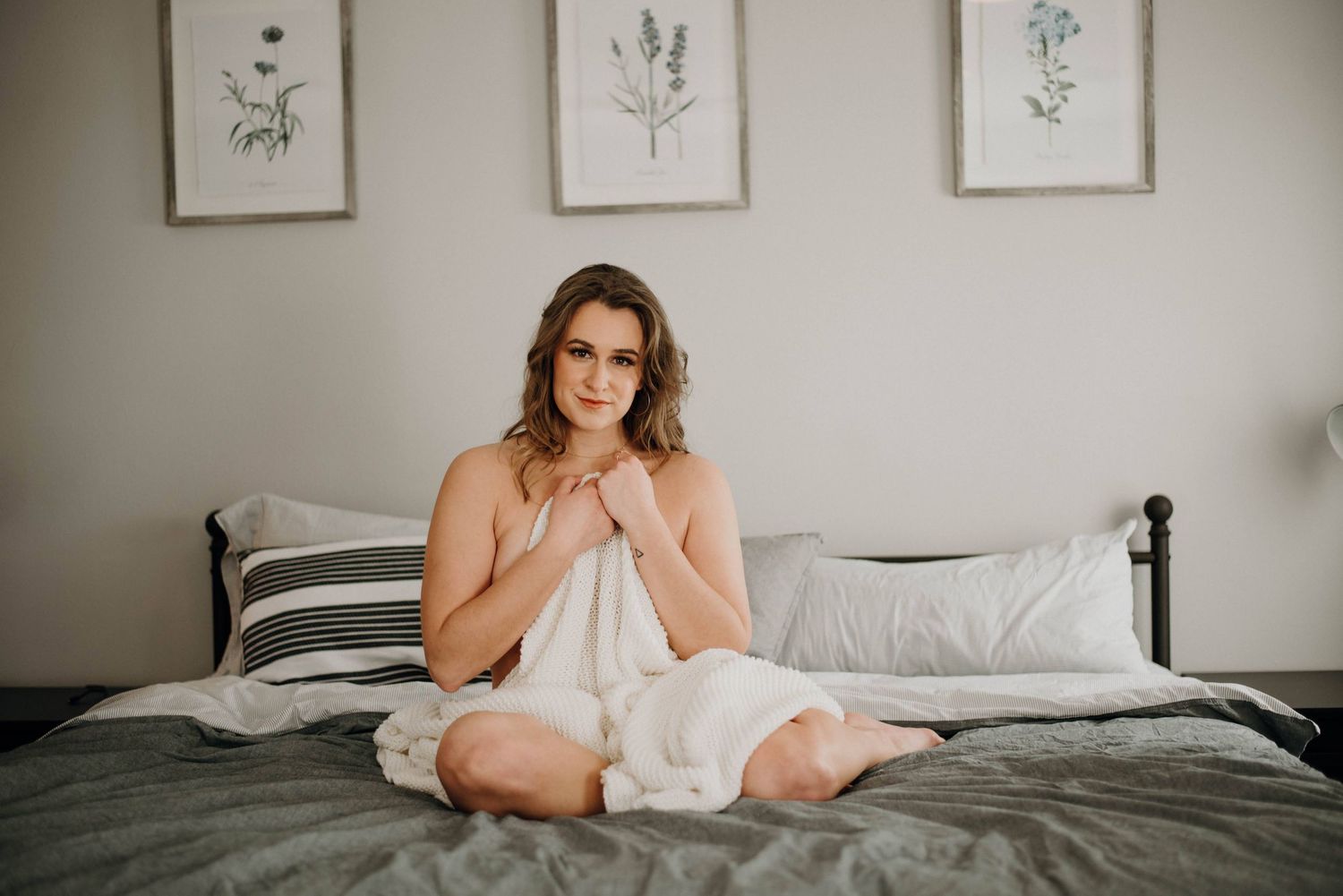 Woman wrapped in white sheet sitting cross-legged on bed with botanical art on wall.