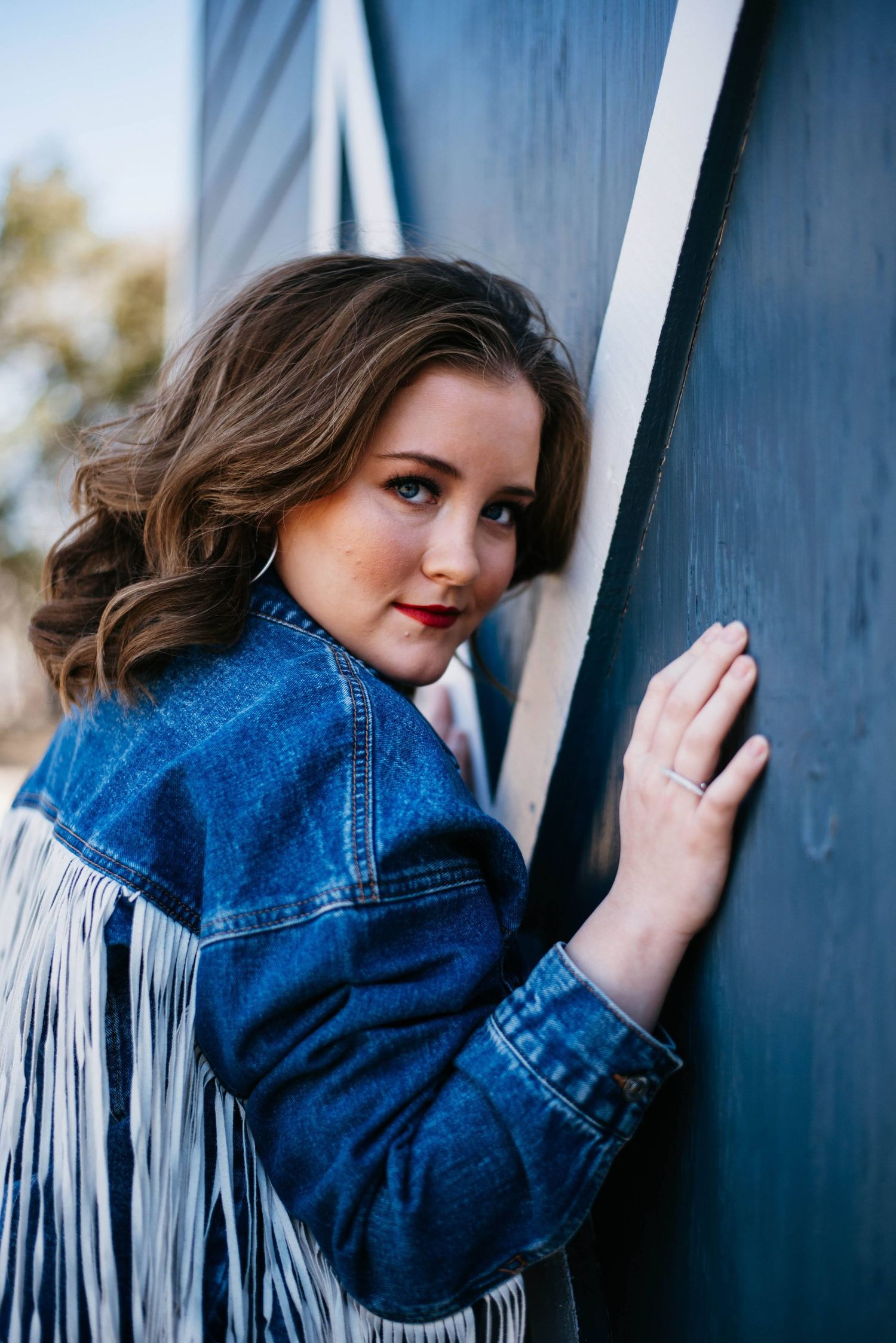 Young woman in fringed denim jacket leaning against a blue wall, looking over her shoulder.