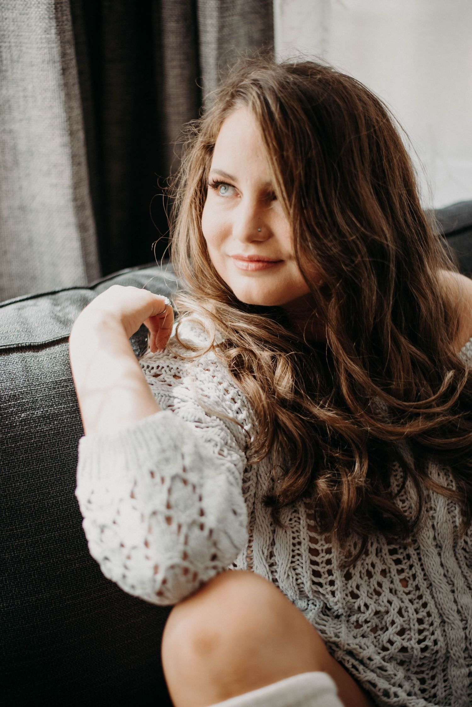 Young woman with long wavy brown hair relaxing on a gray sofa wearing a white knit sweater.