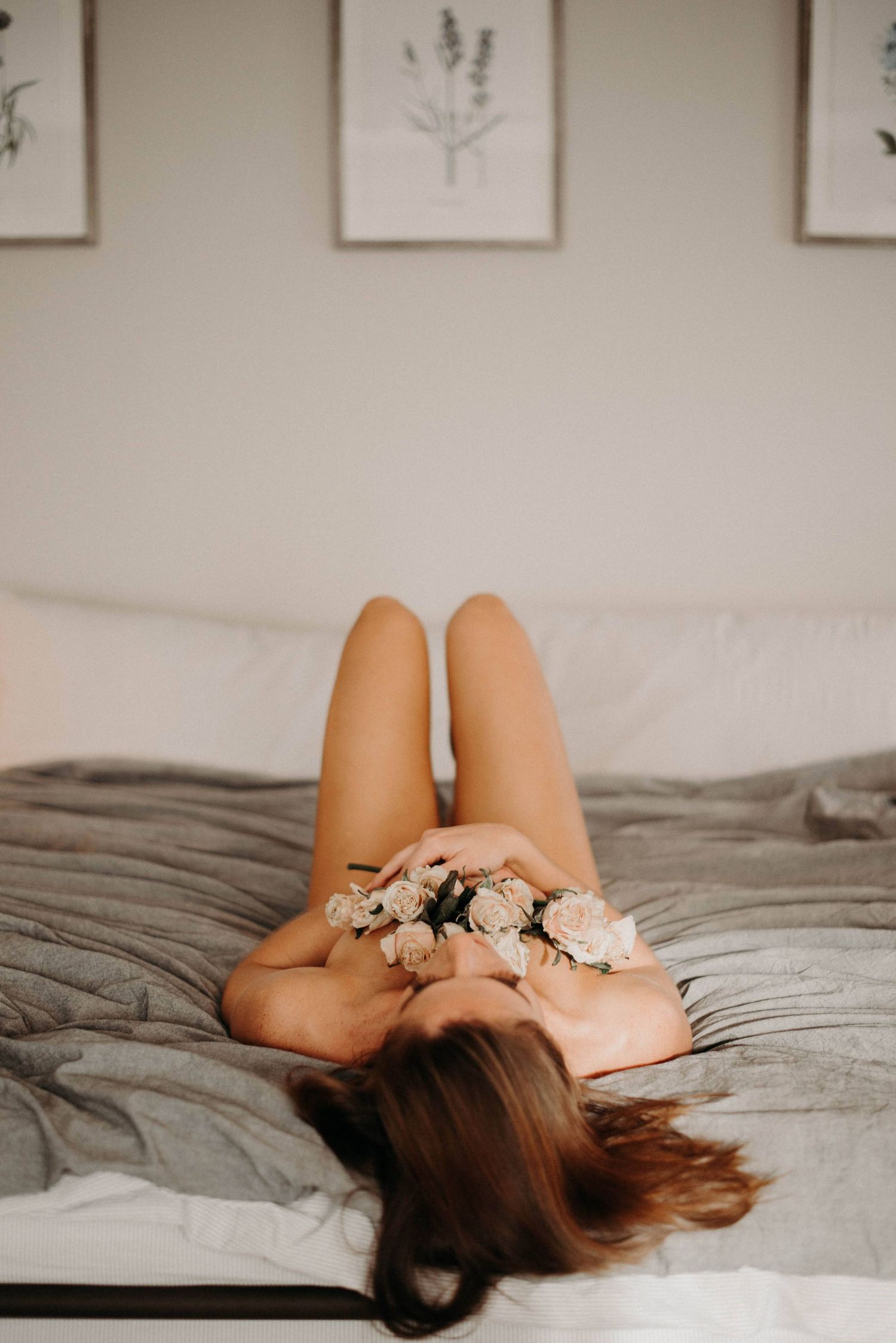 Woman lying on a bed holding white roses with legs raised against a neutral wall.