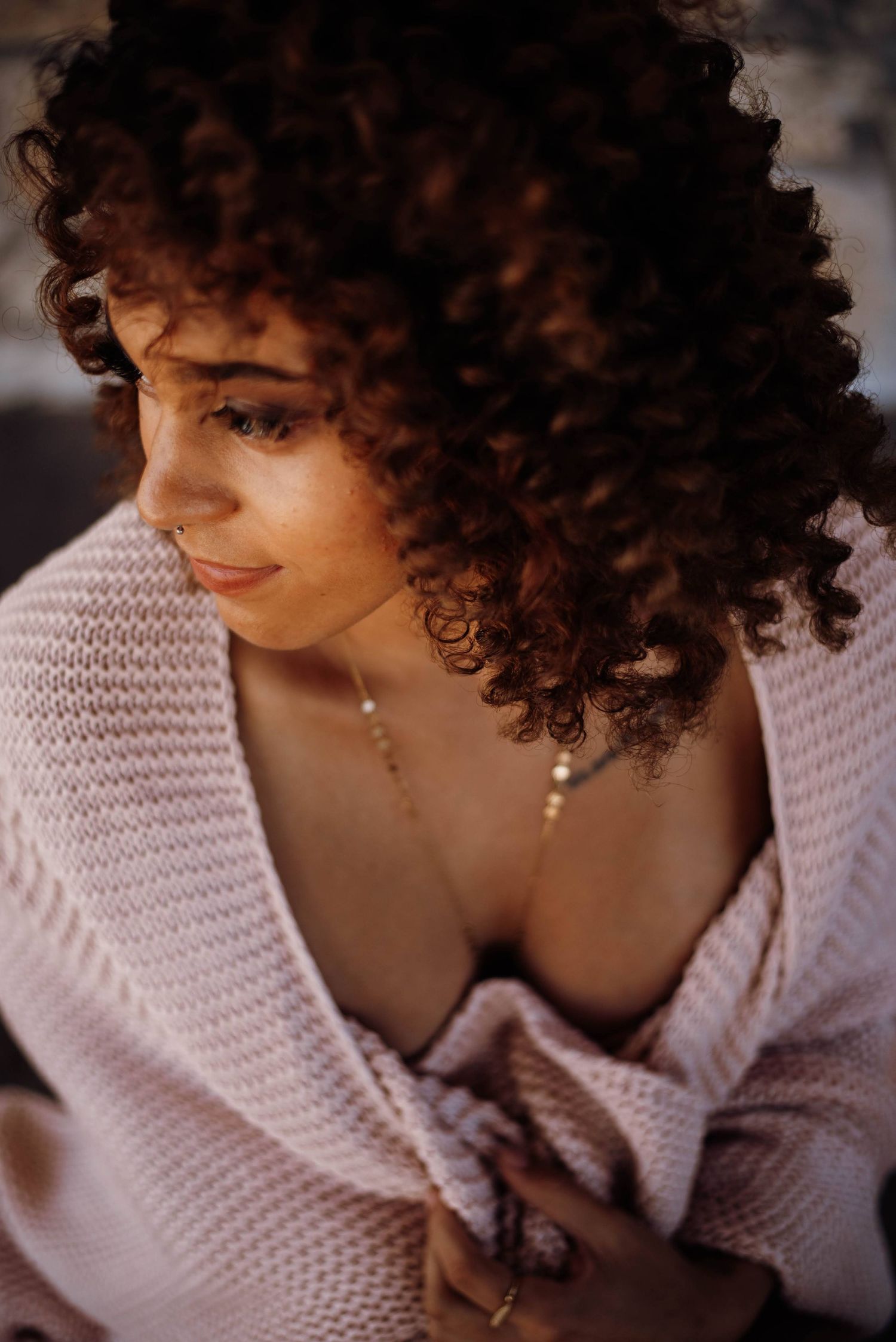 Woman with curly auburn hair wrapped in a pink knit sweater looking down in warm light.