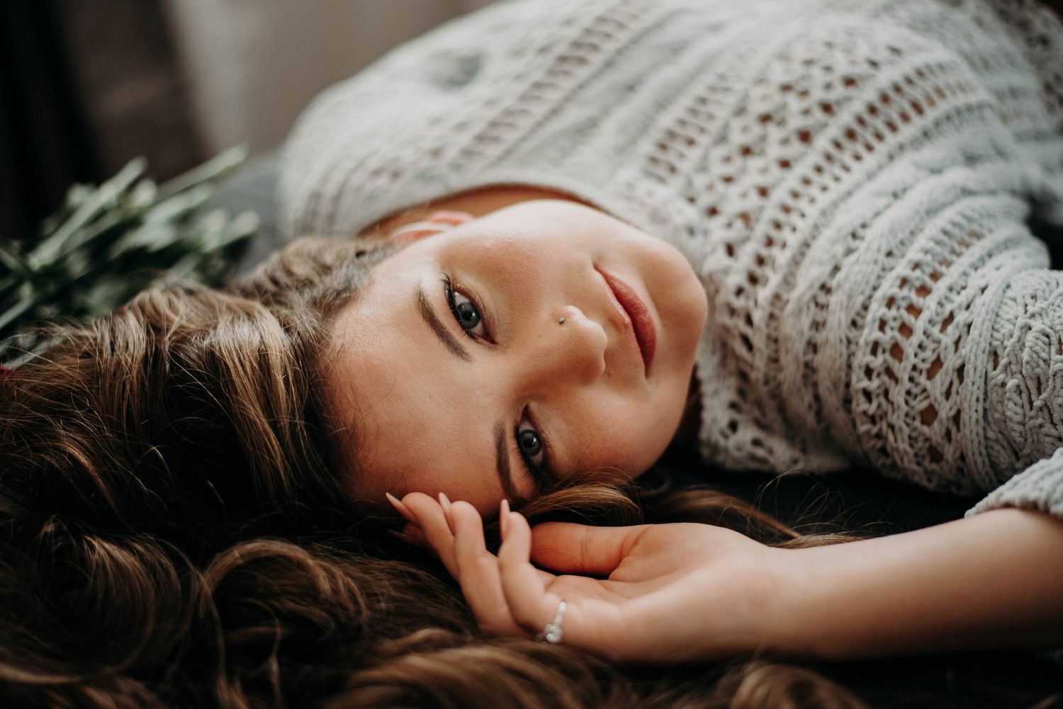 A young woman with long wavy hair lies down wearing a white knit sweater, gazing softly at the camera.