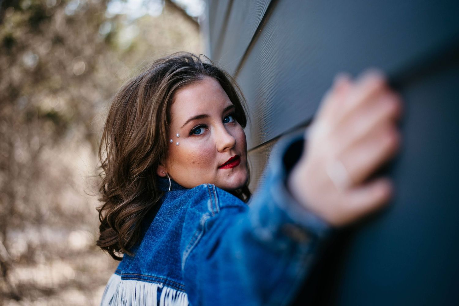 Young woman in denim jacket with red lips leaning against a gray wall outdoors.