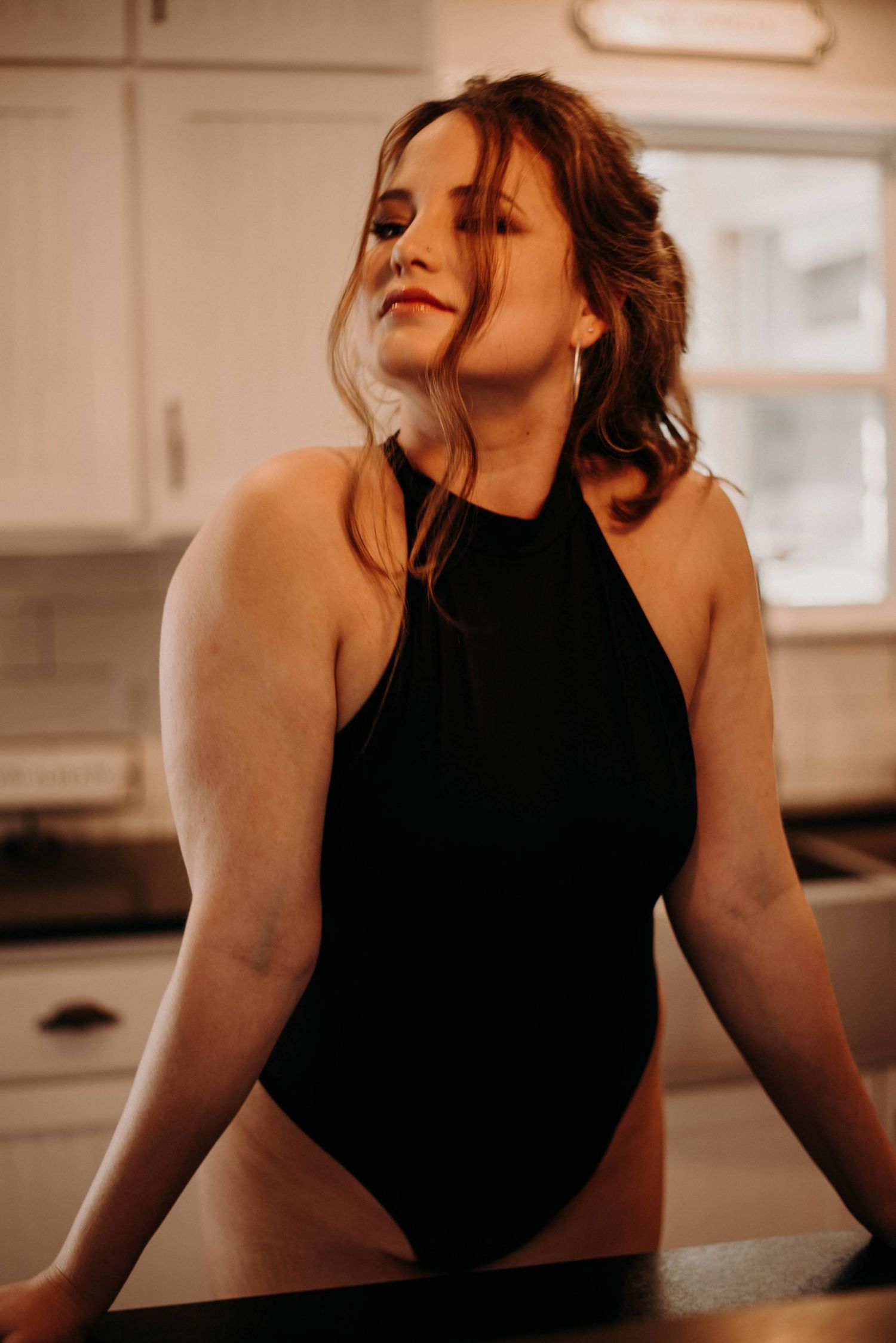 A young woman in a black halter bodysuit leans against a kitchen counter, gazing thoughtfully to the side.