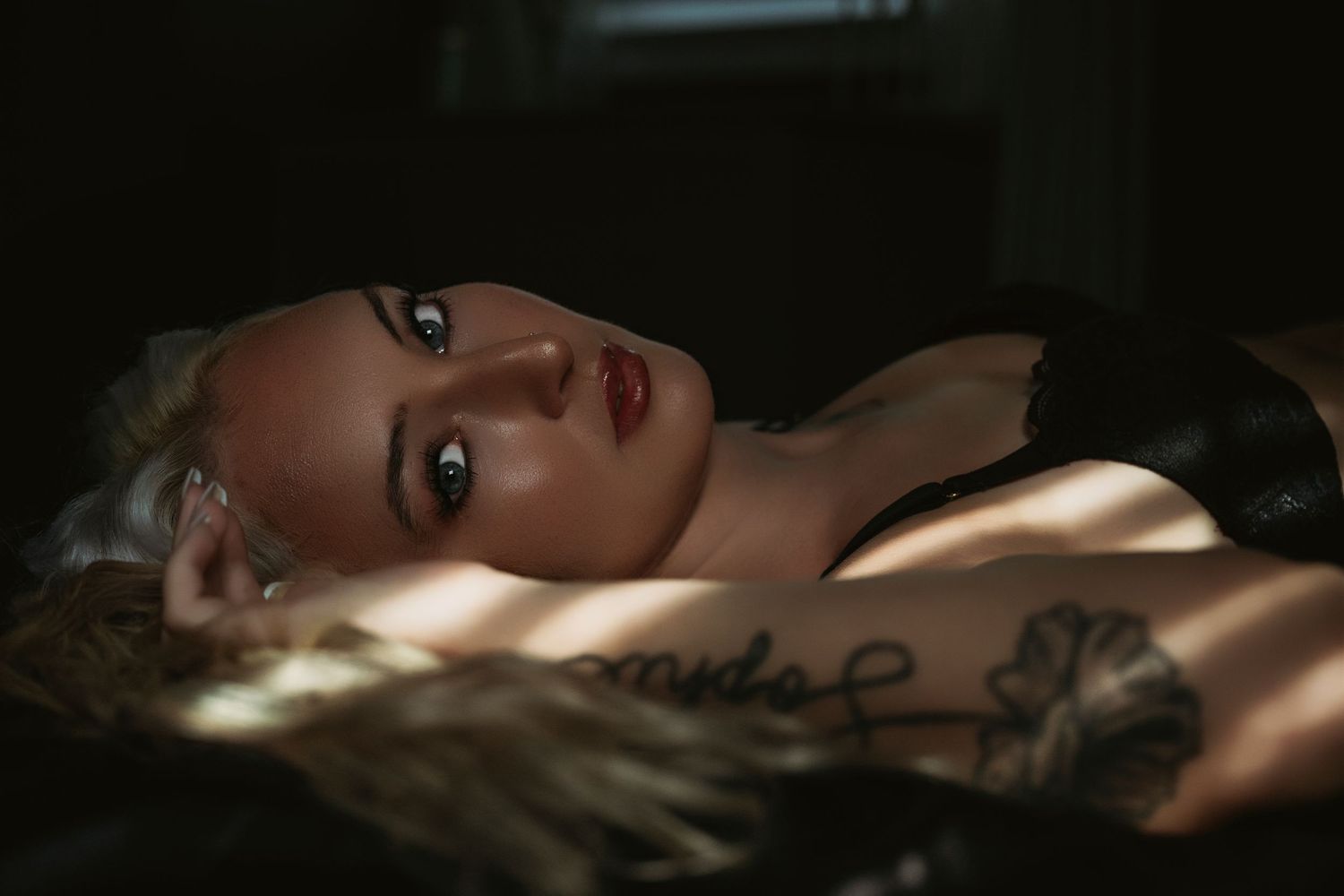 A tattooed blonde woman in black lingerie lying down, gazing upward in dramatic moody lighting.