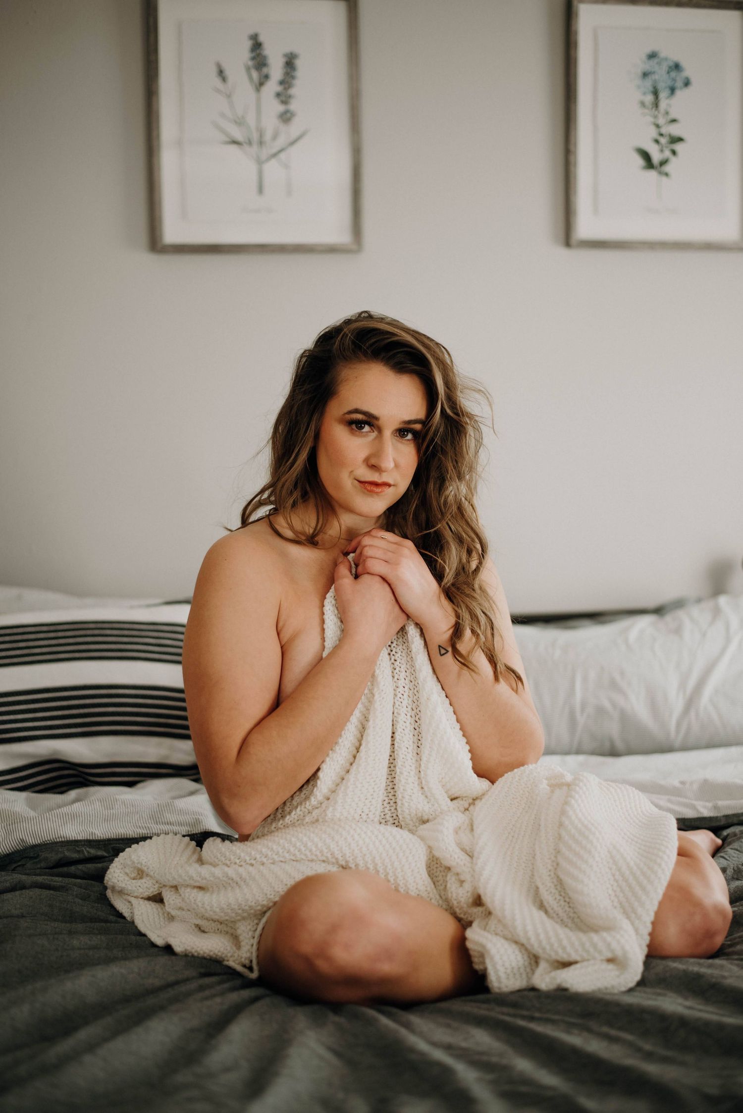 Young woman wrapped in a white knit blanket sitting cross-legged on a bed with botanical art.
