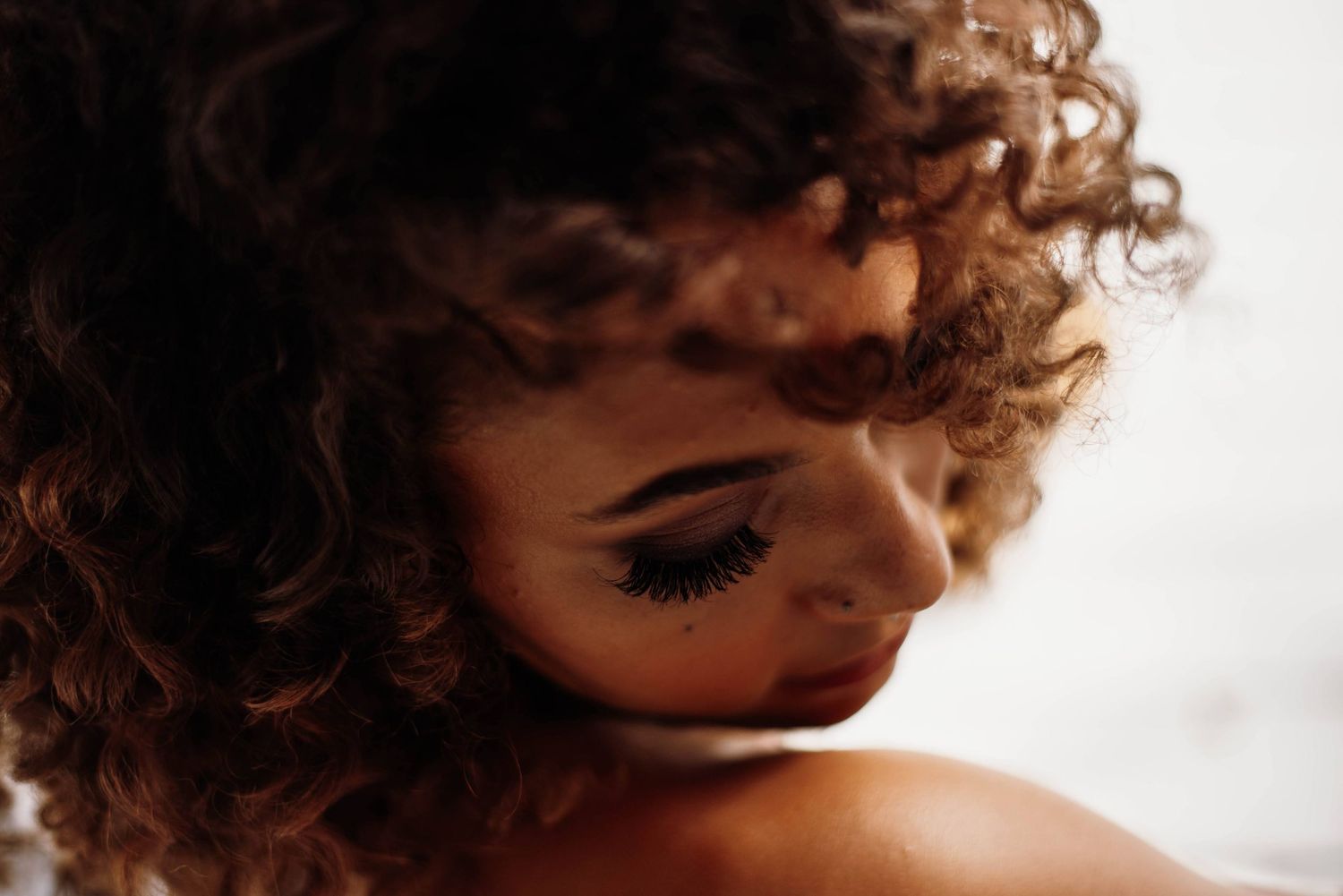 Close-up portrait of woman with curly hair and dramatic lashes, looking down in soft light.