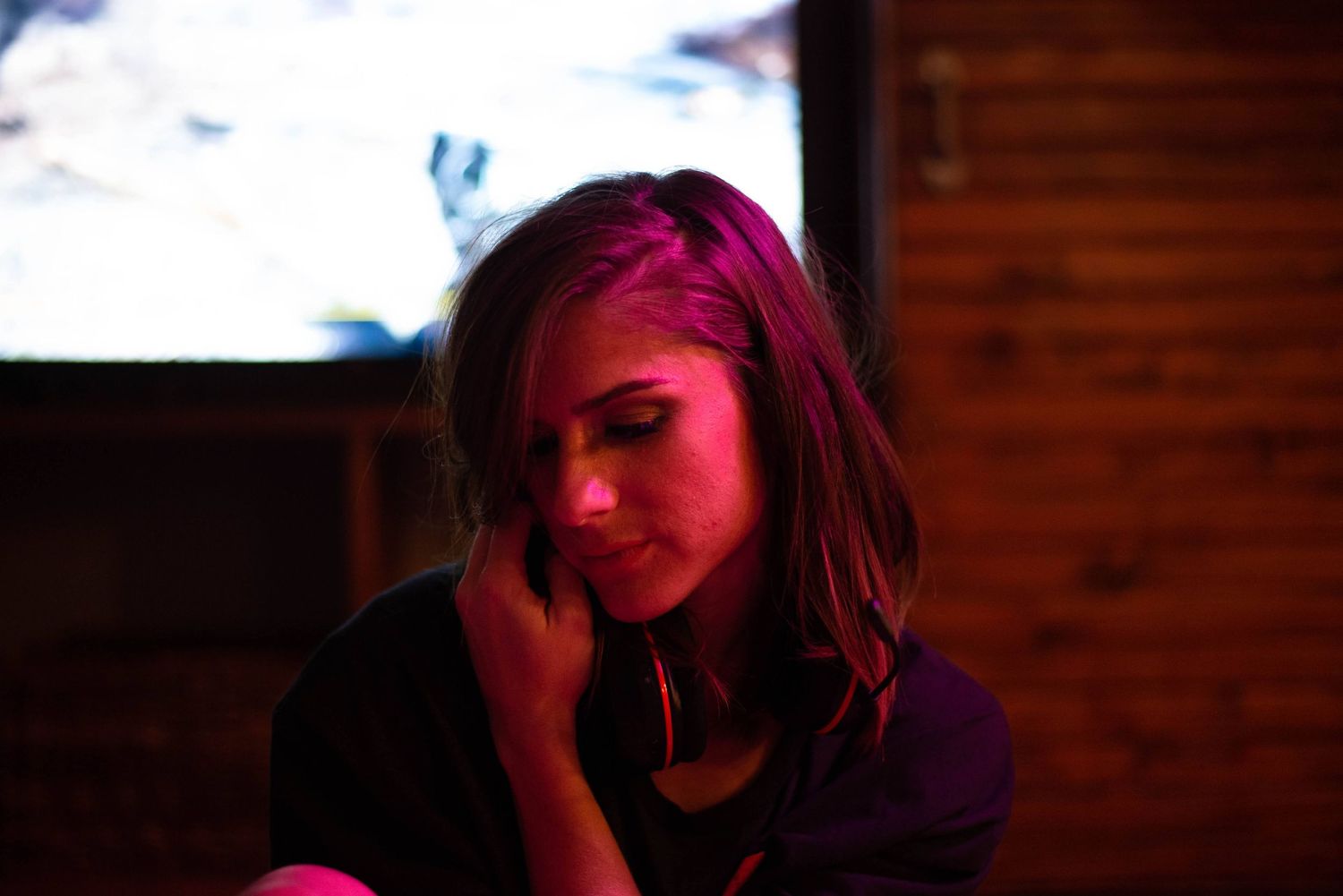 Woman sitting alone in a dimly lit wooden room with soft pink light and a contemplative expression.