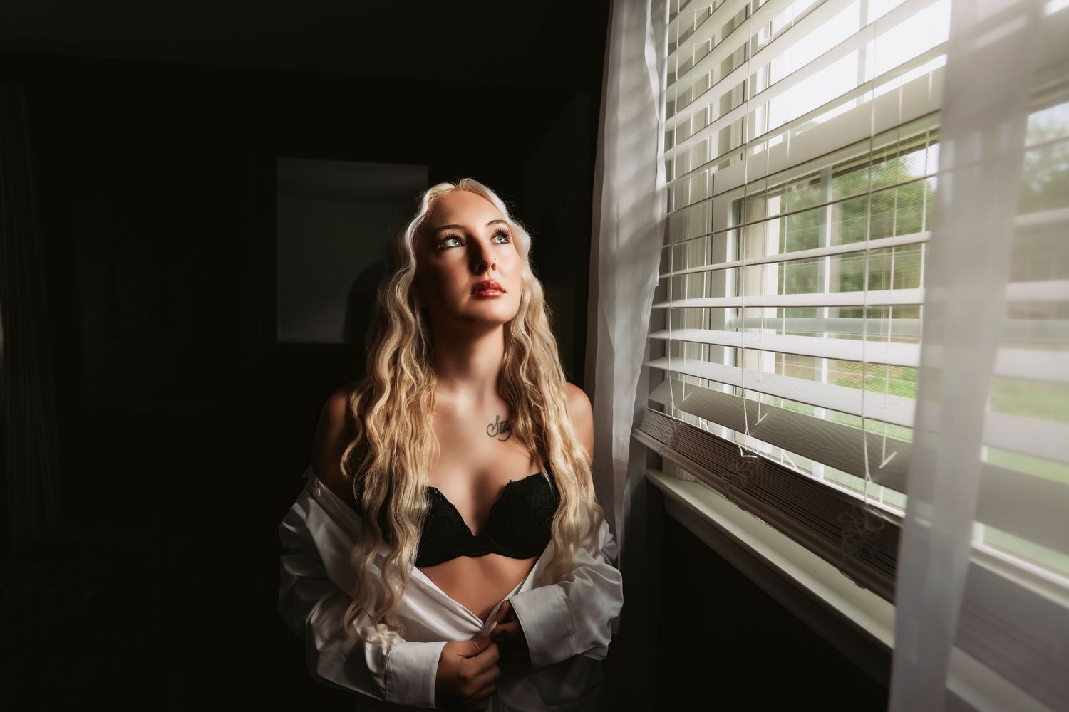 Blonde woman in black bra and open white shirt gazing out window with natural light filtering through blinds.