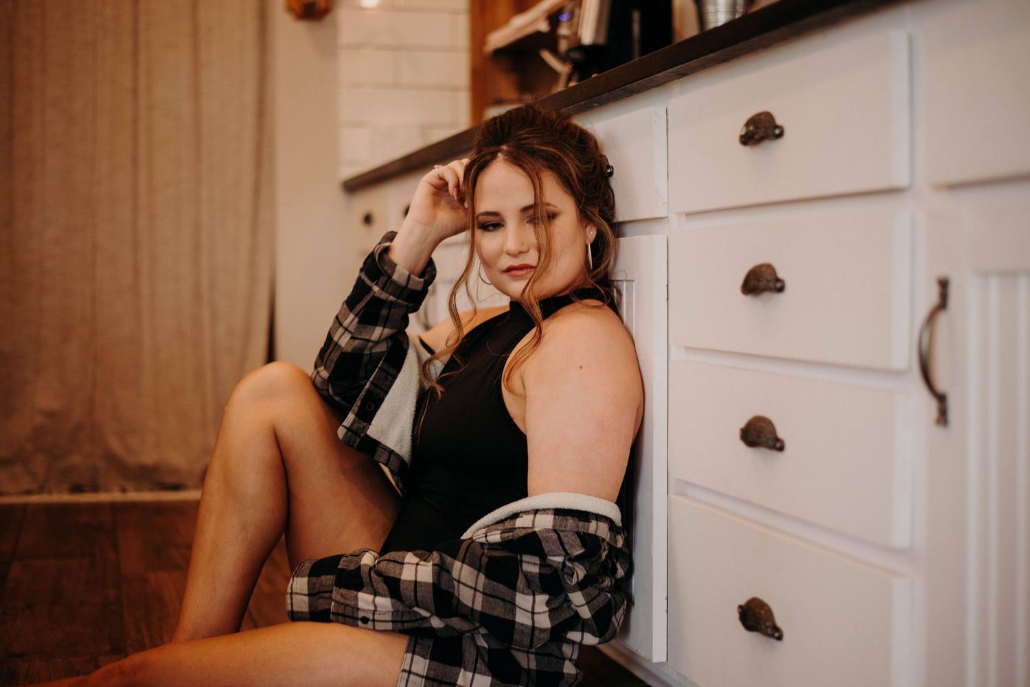 Young woman in plaid shirt and black top sitting casually on kitchen floor leaning against white drawers.