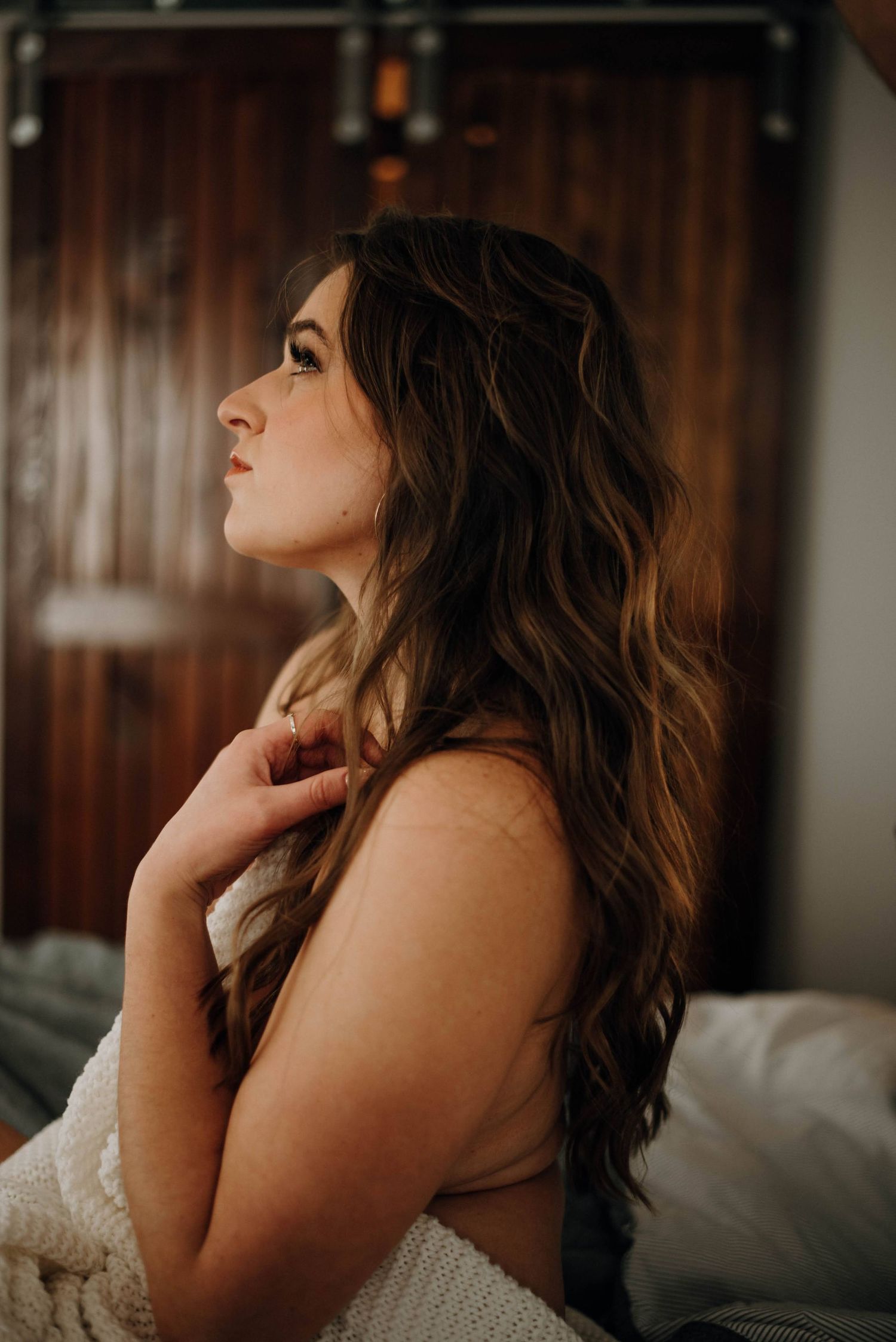 A woman with long wavy brown hair sits on a bed wrapped in a white blanket, gazing upward in soft moody lighting.
