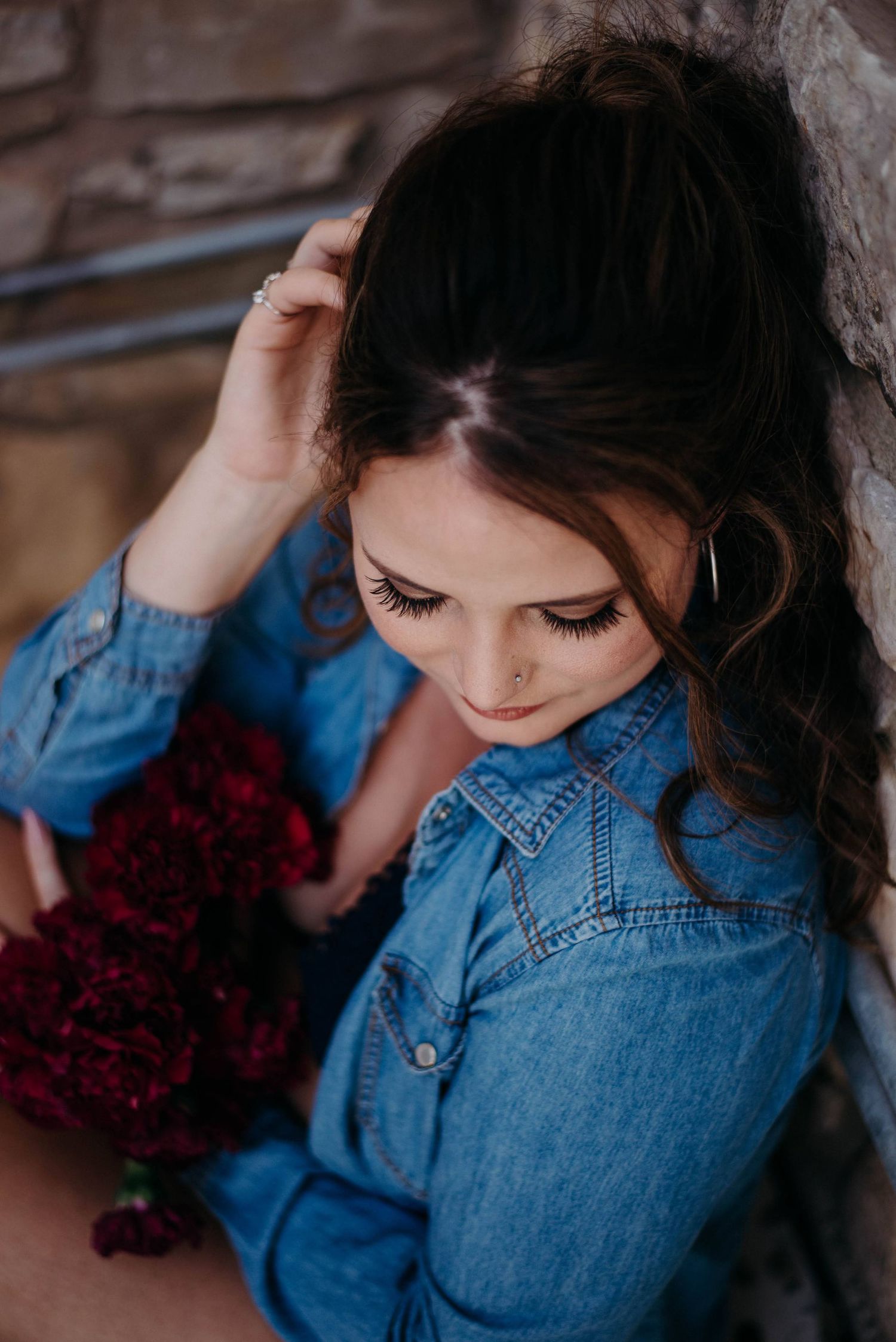 Young woman in denim shirt holding red flowers against a stone wall, looking down thoughtfully.