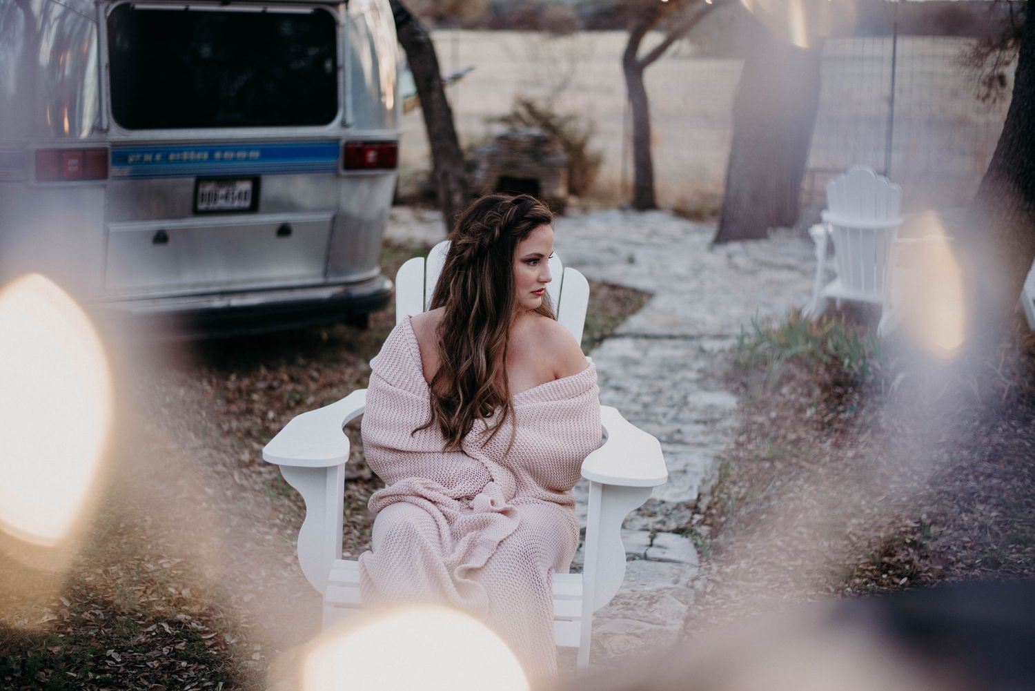 Woman in pink off-shoulder dress sitting in white chair outdoors near vintage van in soft bokeh light.