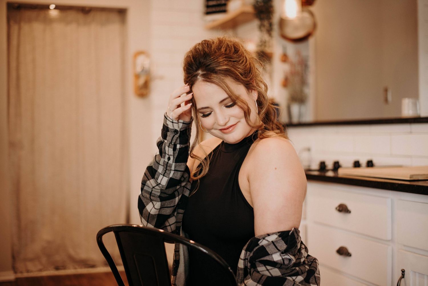 Young woman in black top and plaid shirt smiling with eyes closed in a cozy kitchen setting.