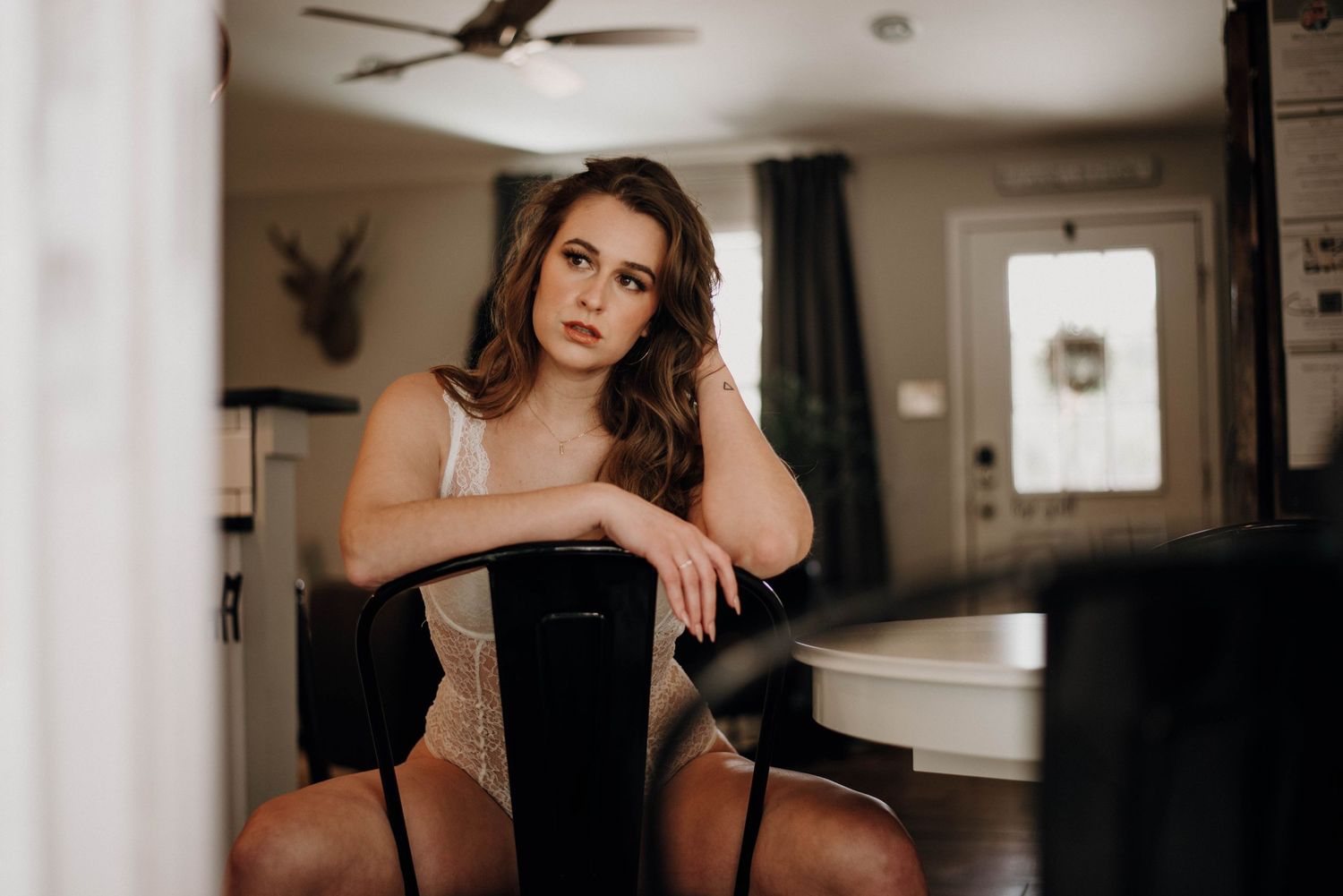 A young woman in lingerie sits pensively on a chair in a softly lit room, gazing into the distance.